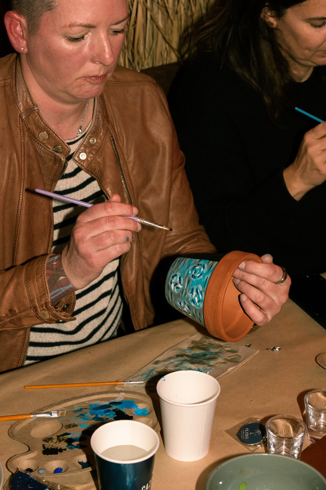 Two women sitting at a table, one holding a small paintbrush and a painted flowerpot, with various painting supplies and cups on the table.