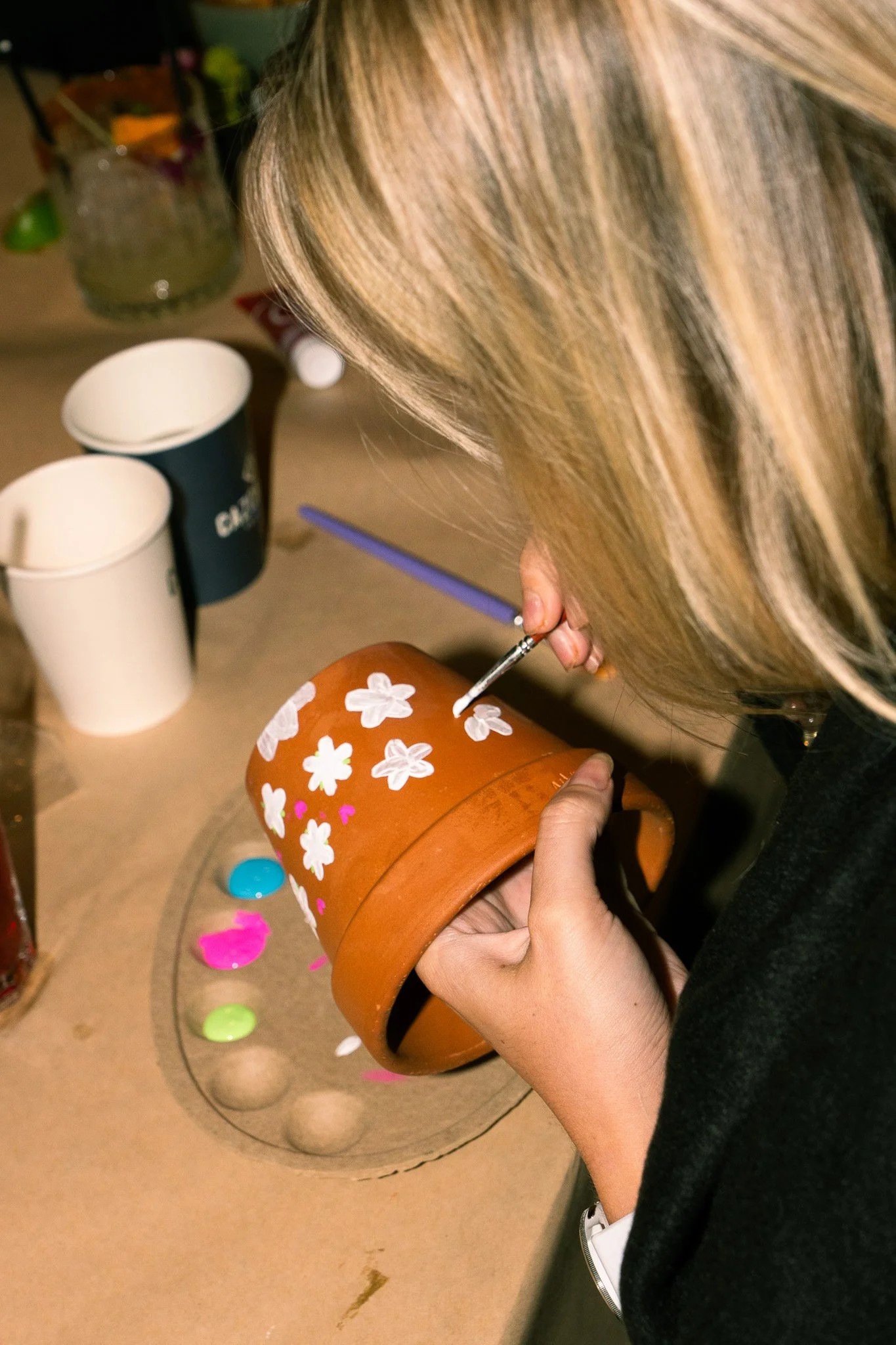 Person painting white flowers on a brown flower pot, with various paints and cups nearby.