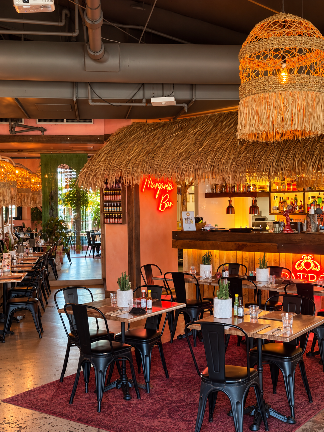 Indoor restaurant with black chairs, wooden tables with salt and pepper shakers, small potted cacti, and water glasses. Decor includes thatched roof accents, neon signs for Margarita Bar and patio seating, and warm ambient lighting.