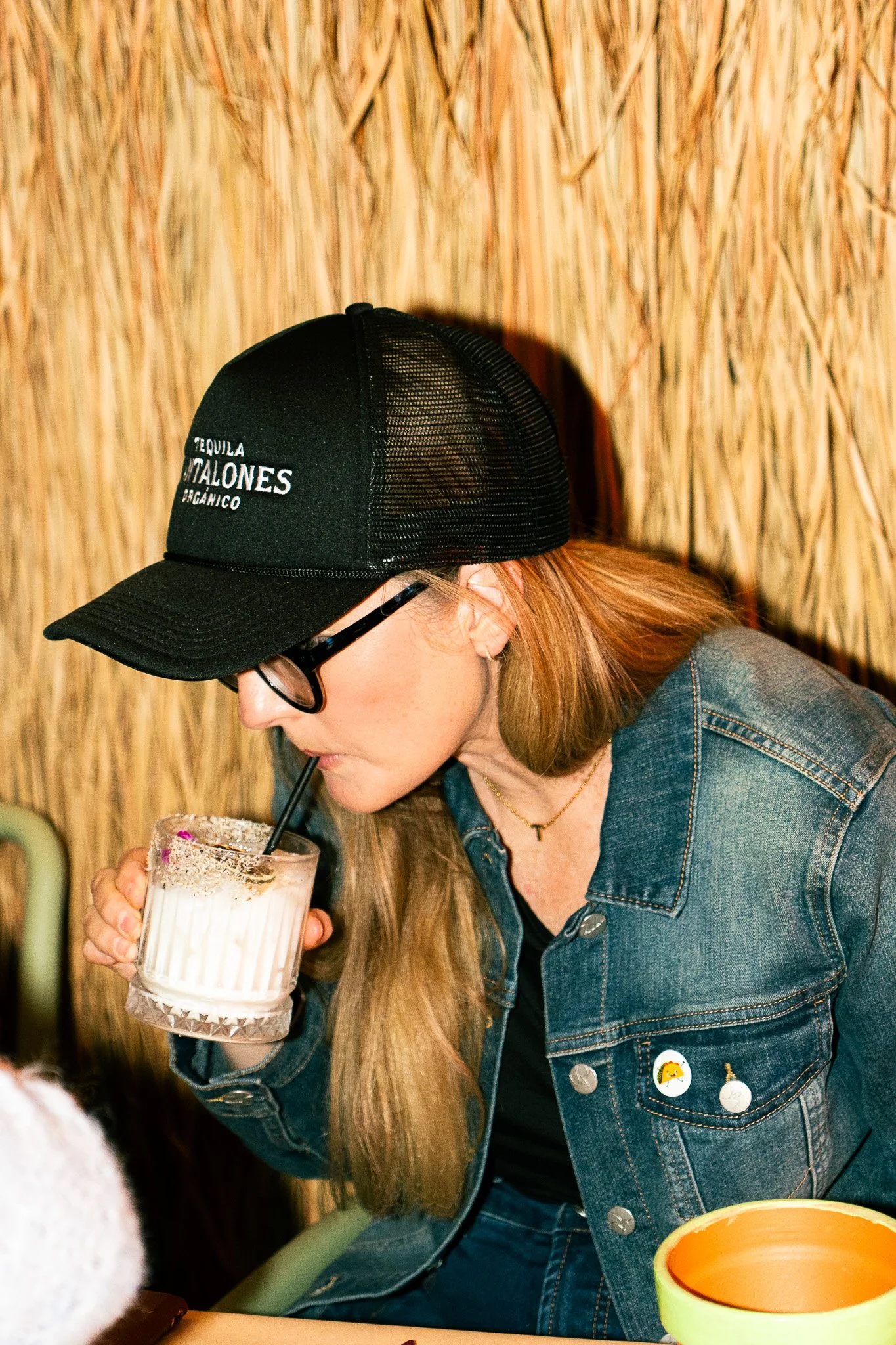 A woman with long hair, wearing glasses, a denim jacket with decorative pins, and a black shirt, is drinking a beverage with a straw from a glass mug. She is in front of a bamboo wall.