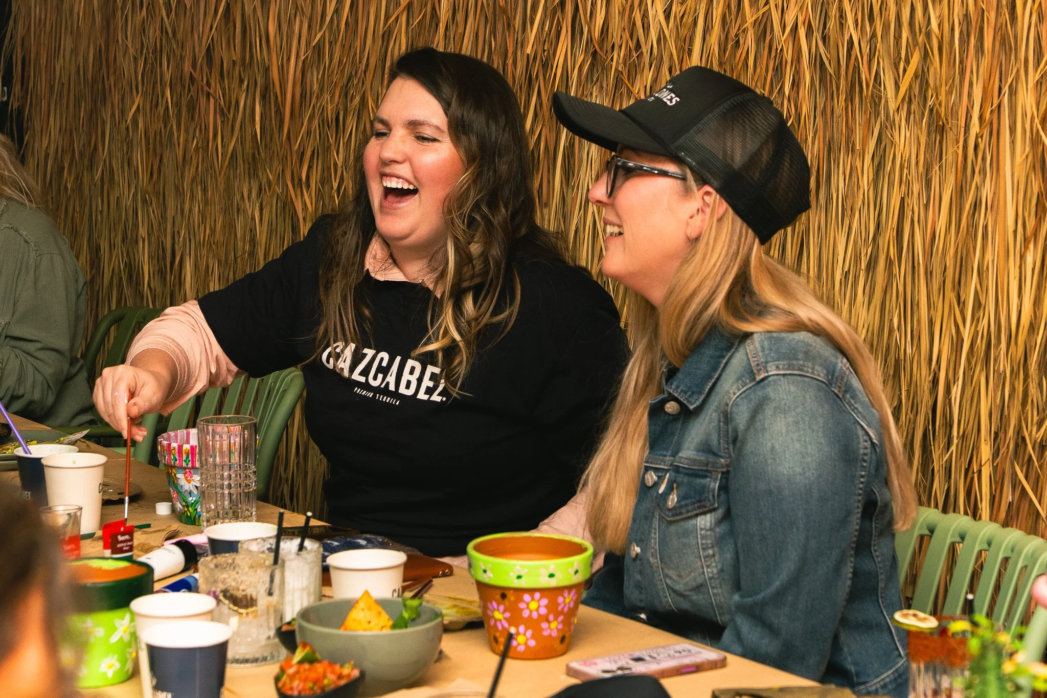 Women laughing and talking at a party or gathering, sitting at a table with colorful cups and food, in a setting with straw or grass-like decor.