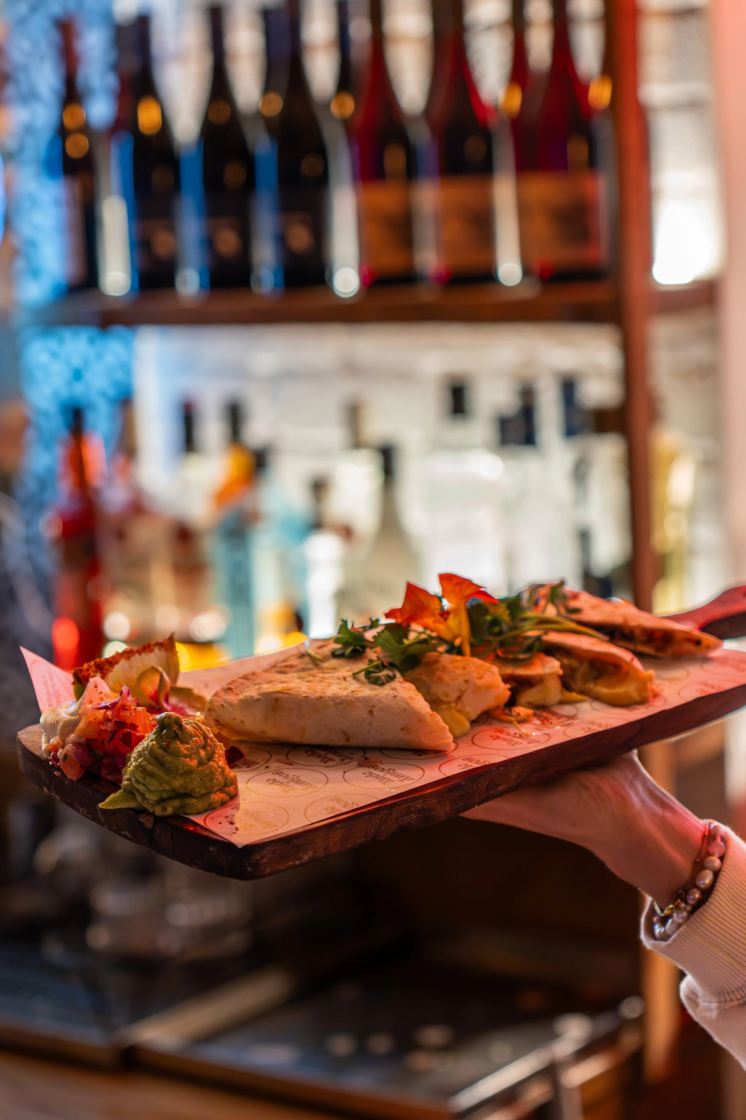 A hand holding a wooden tray with Mexican food, including tacos, guacamole, and salsa, at a restaurant with bottles on shelves in the background.