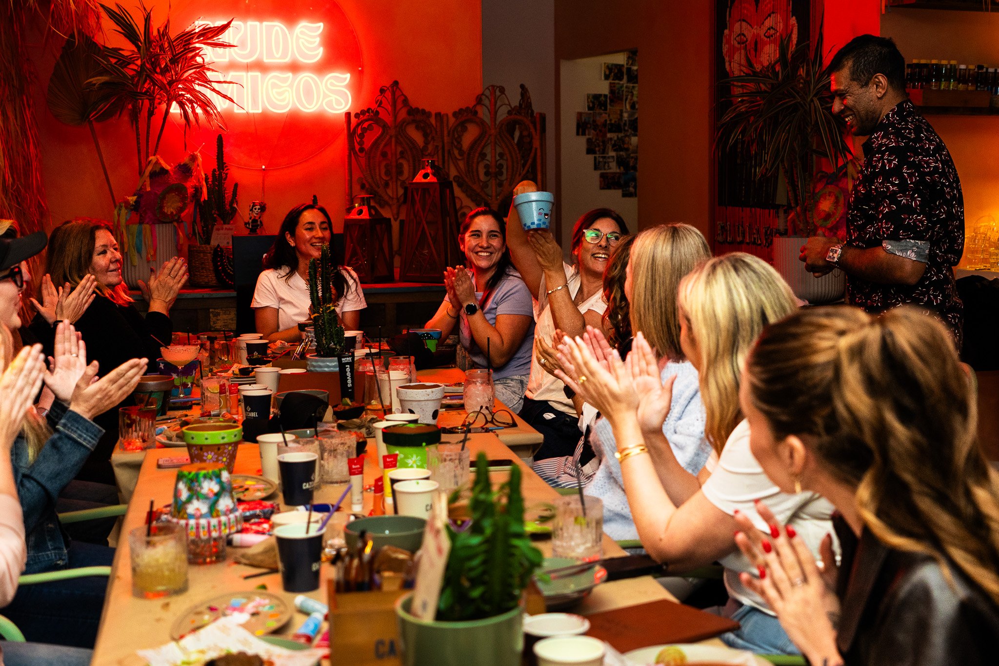 A group of people at a party sitting around a long table, clapping and smiling. There is a man standing and smiling at the right, and a woman holding a cup in the middle. The table is decorated with party supplies and snacks. The background has a red neon sign and tropical plants, creating a lively festive atmosphere.