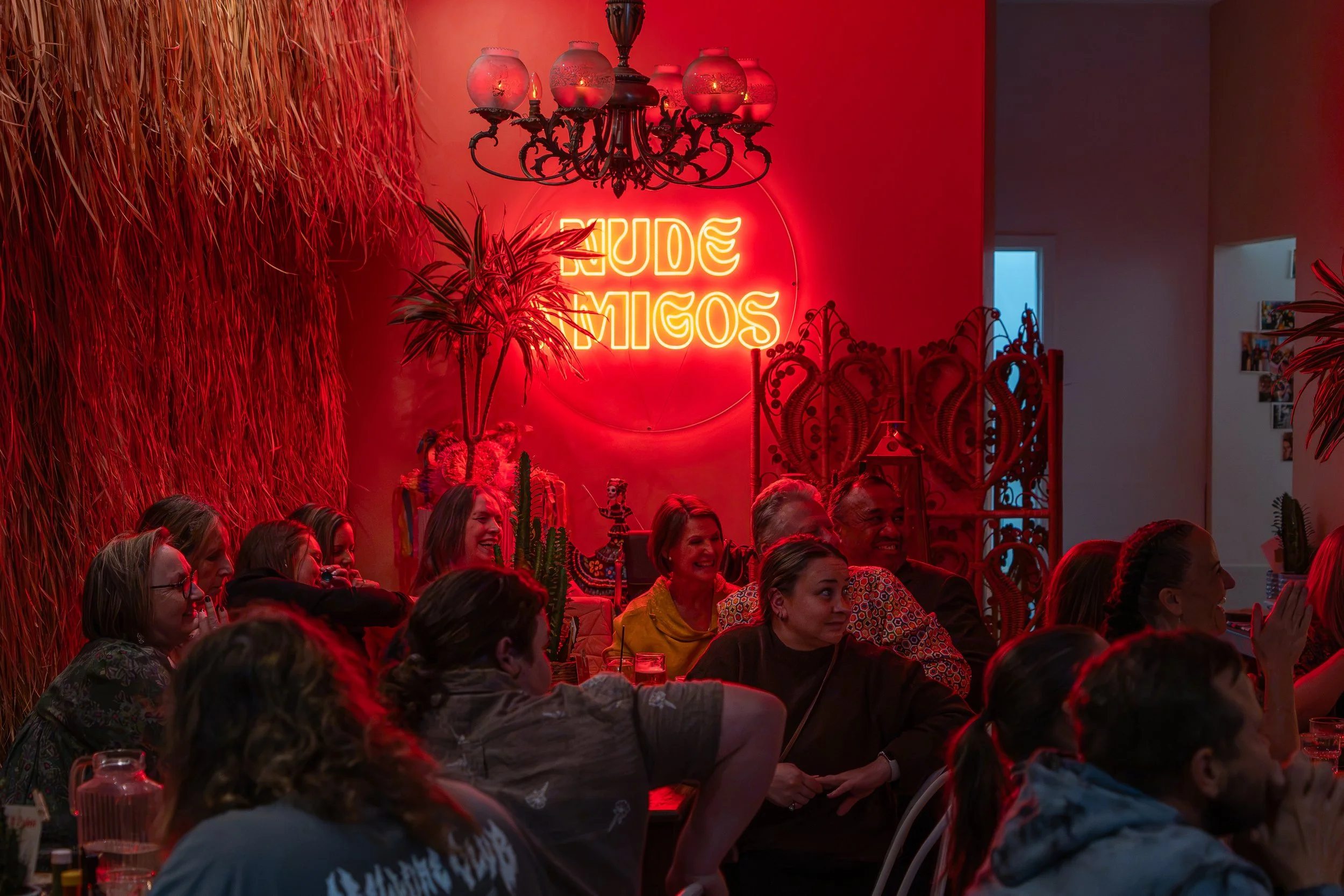 People sitting at a restaurant or party with a red neon sign that says 'California.' Red lighting, decorative plants, a chandelier, and vibrant decor are visible.