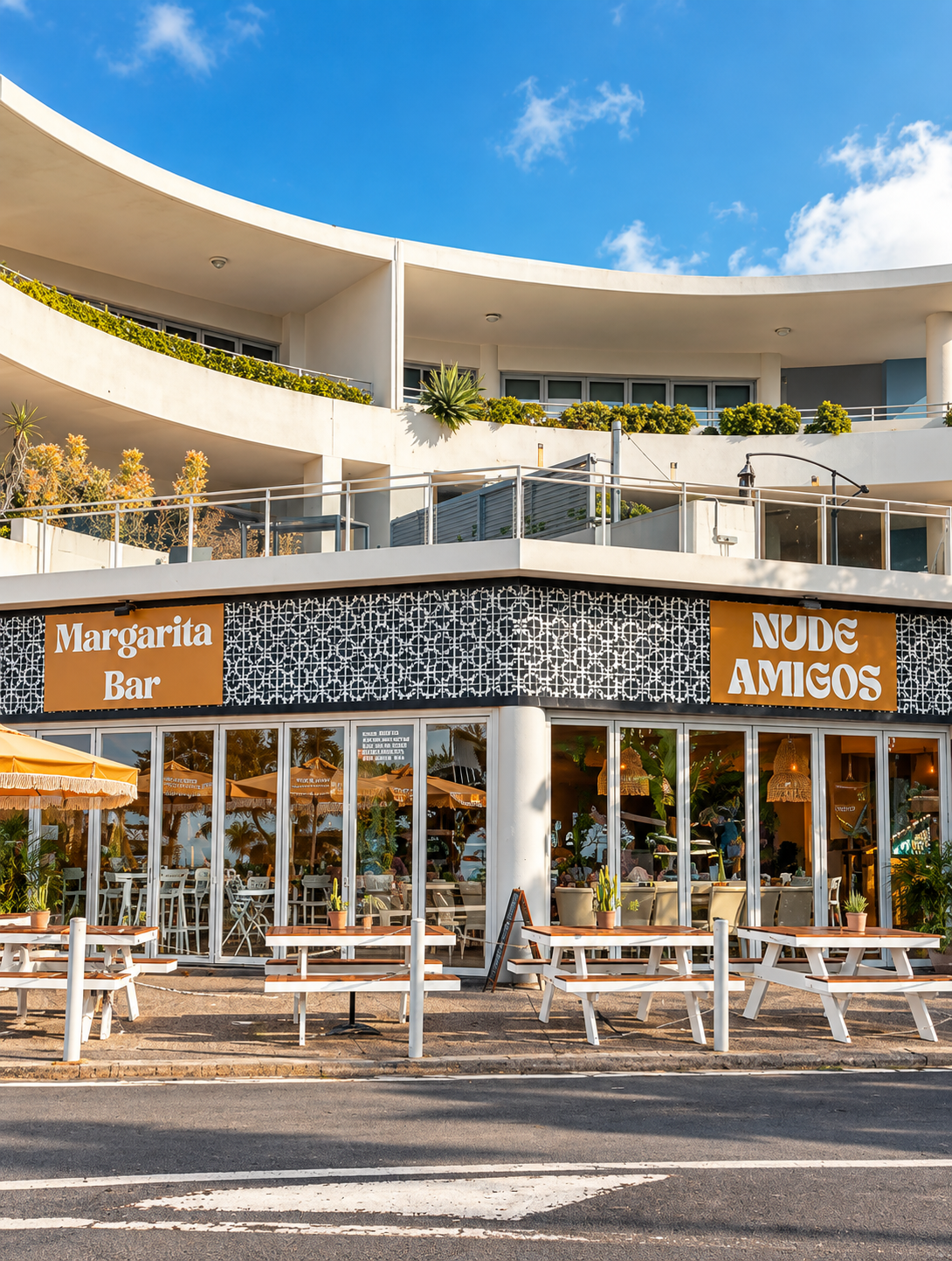 Exterior of a restaurant with outdoor seating and a sign that reads 'Margarita Bar' and 'Nude Amigos'. The restaurant has large glass windows, white and black patterned walls, and potted plants outside. The building has multiple stories with balconie