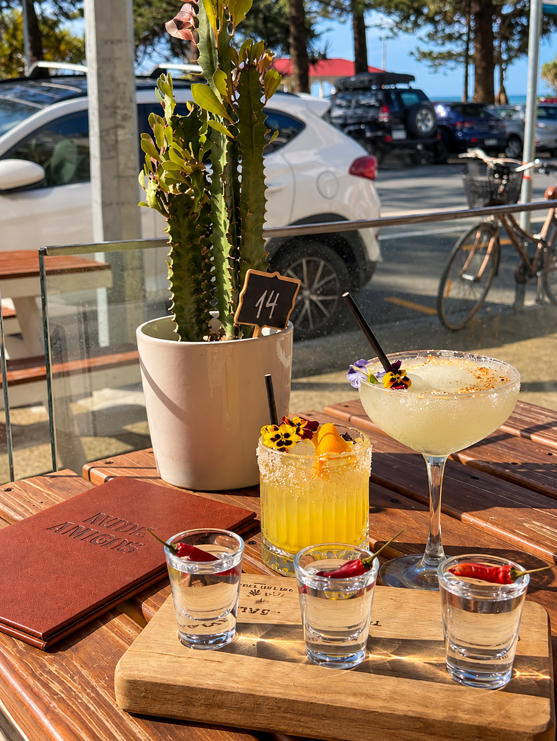 A wooden table displays two cocktails with flowers and garnishes, shot glasses with cherries, a menu, and a potted cactus. Outside the window, cars, a bicycle, and trees are visible.