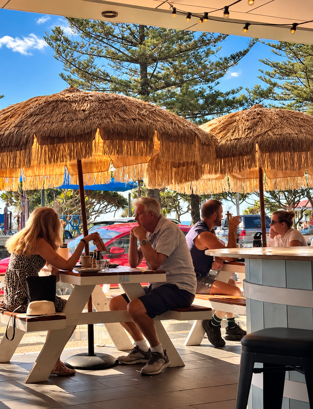People sitting at a picnic table under straw umbrellas outside at a cafe or restaurant on a sunny day, with trees and parked cars in the background.
