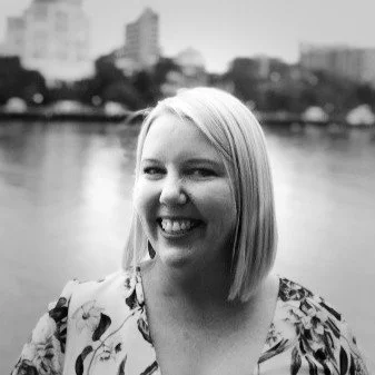 A smiling woman with shoulder-length blonde hair standing outdoors near a river with city buildings in the background.