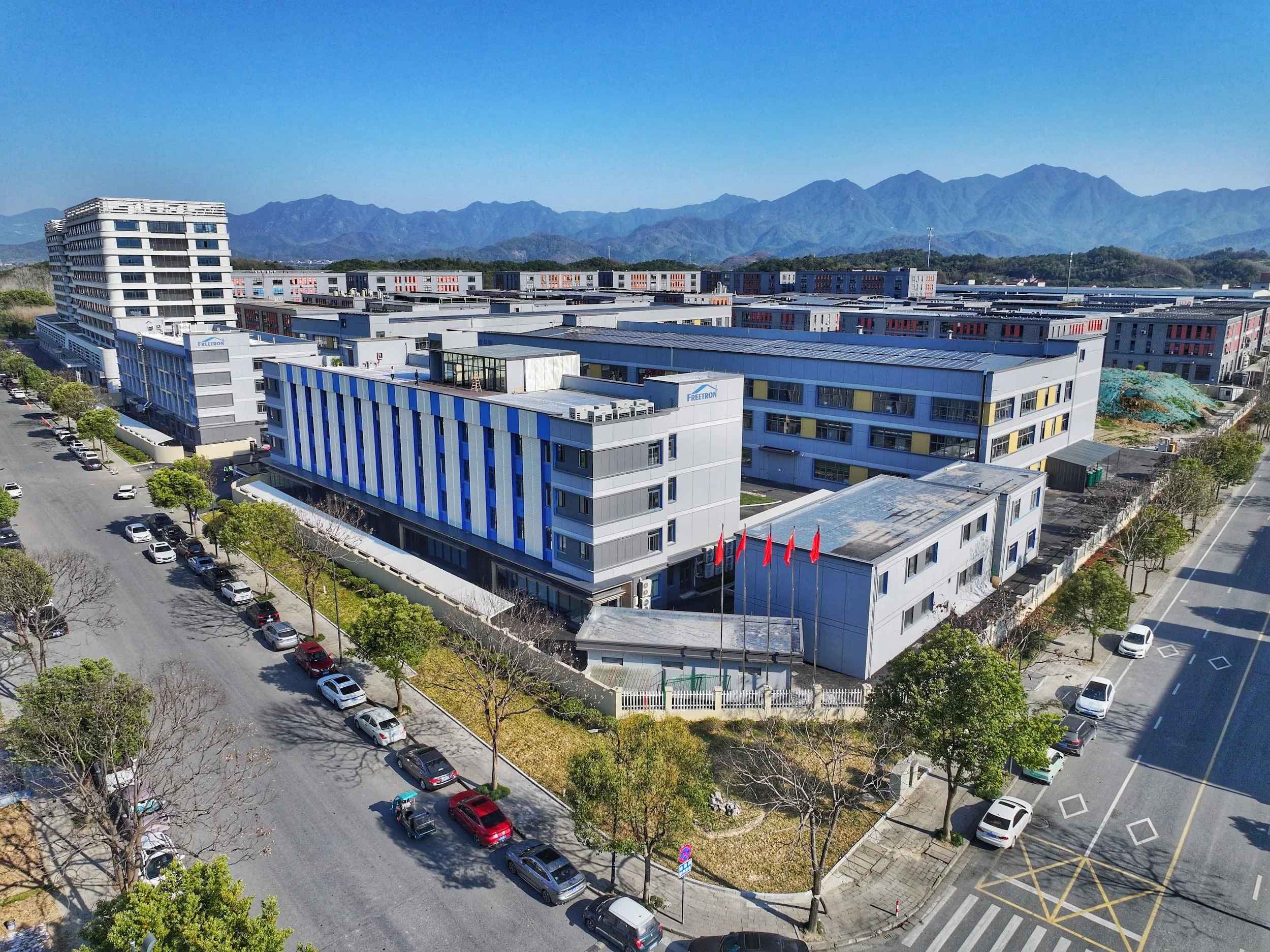 Aerial view of a modern, multi-story building complex with surrounding parking lot and trees, set against a backdrop of mountain range under a clear blue sky.