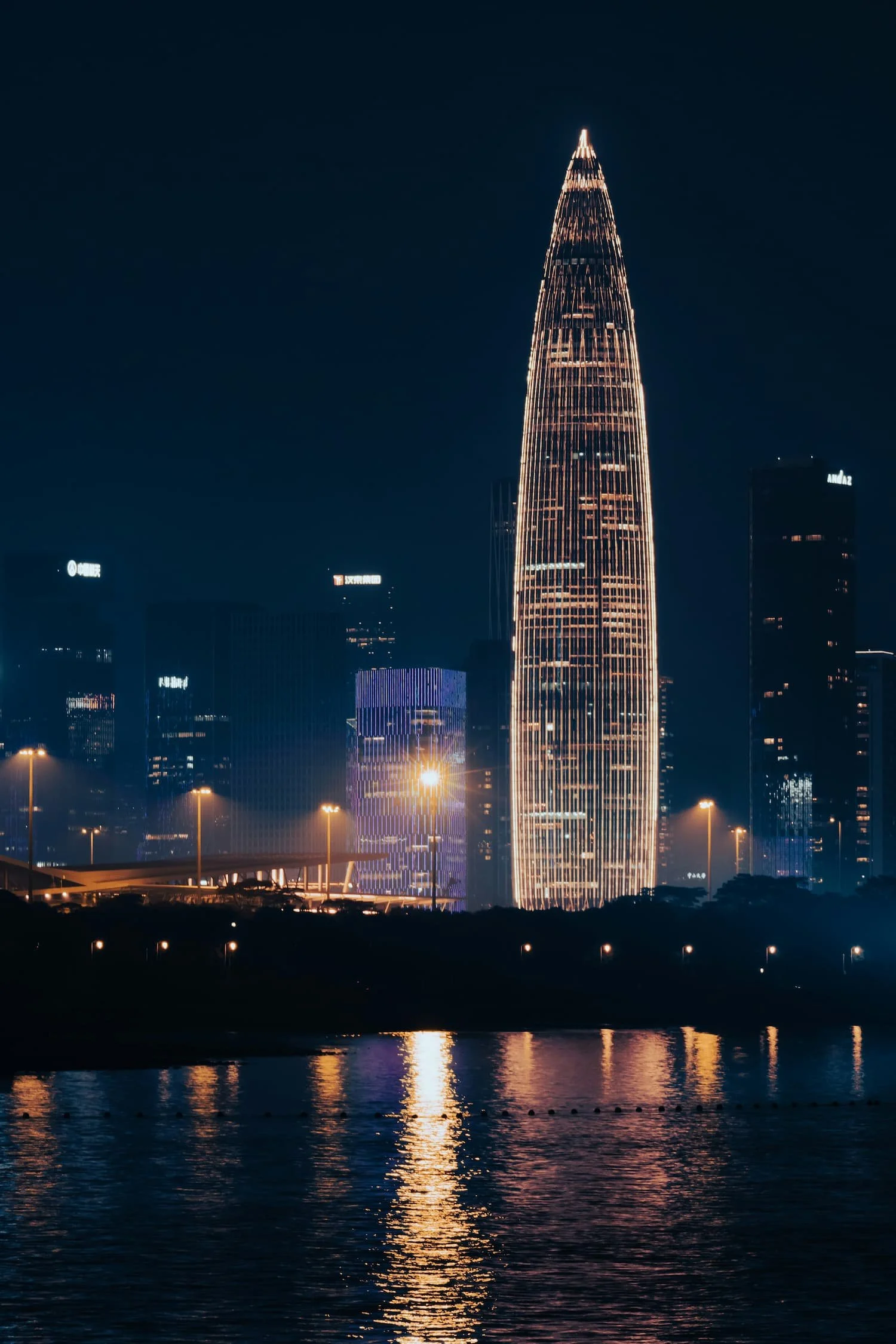Nighttime cityscape featuring a tall, illuminated skyscraper with vertical light lines, reflections on a body of water, and surrounding buildings with lights.