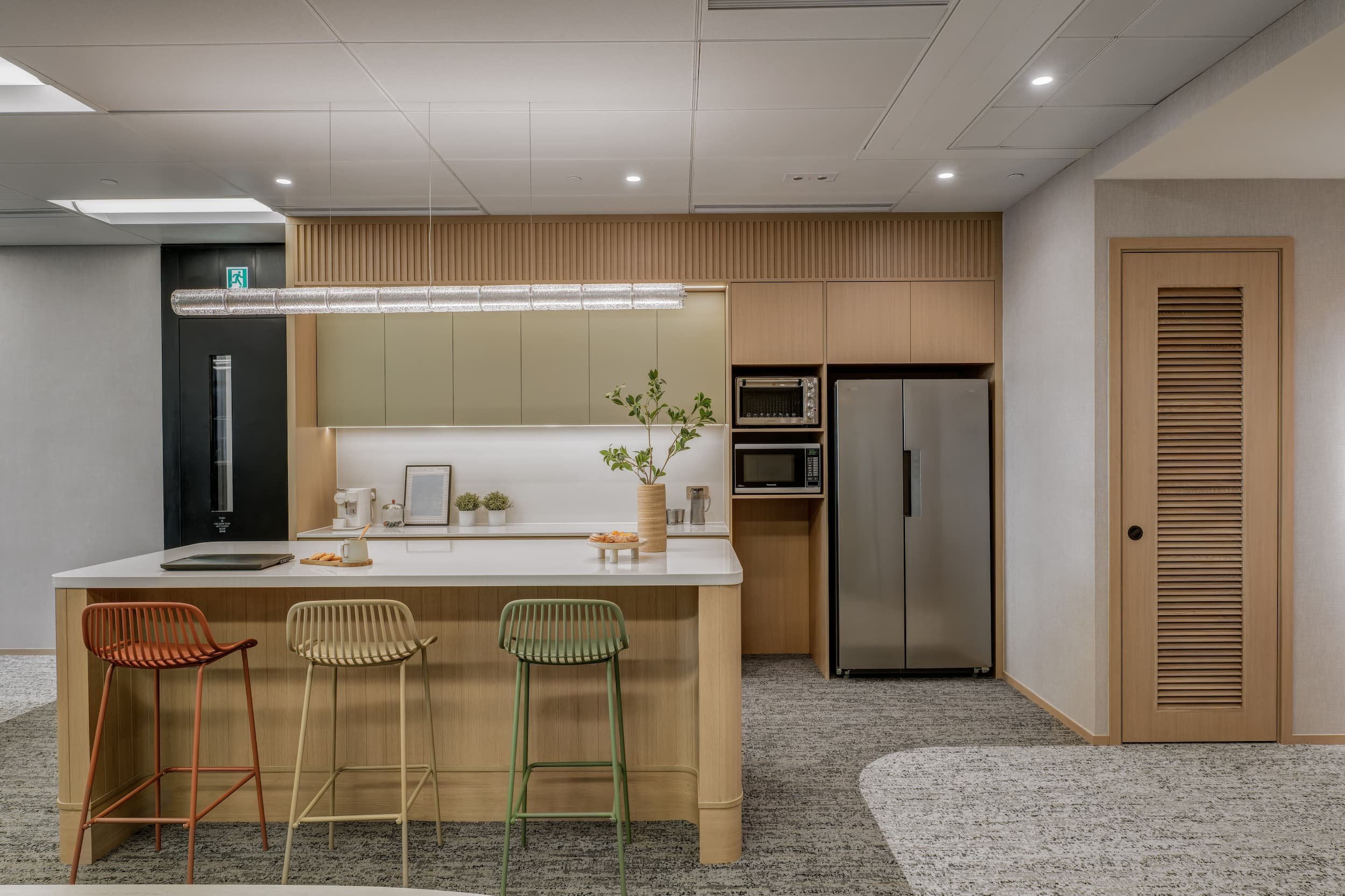 Modern kitchen with light wood cabinets, white countertop, and a small potted plant. There are colorful chairs at a breakfast bar and built-in appliances including a microwave and refrigerator. The ceiling has recessed lighting and a duct system.