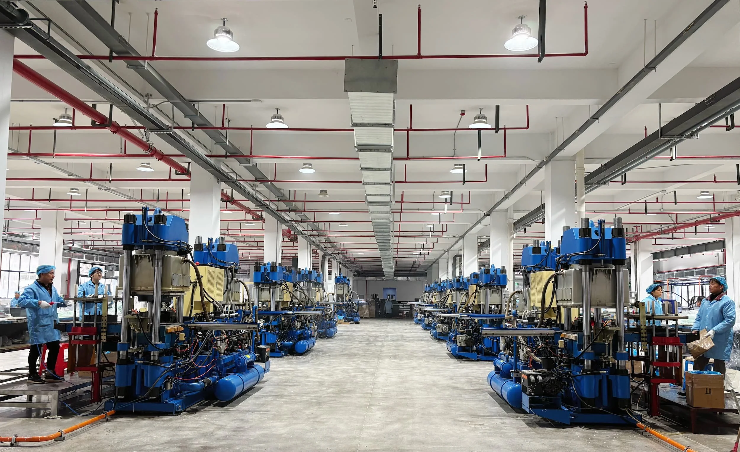 Industrial manufacturing facility with workers in blue uniforms and hair coverings operating machinery, large pipes, and overhead lighting.