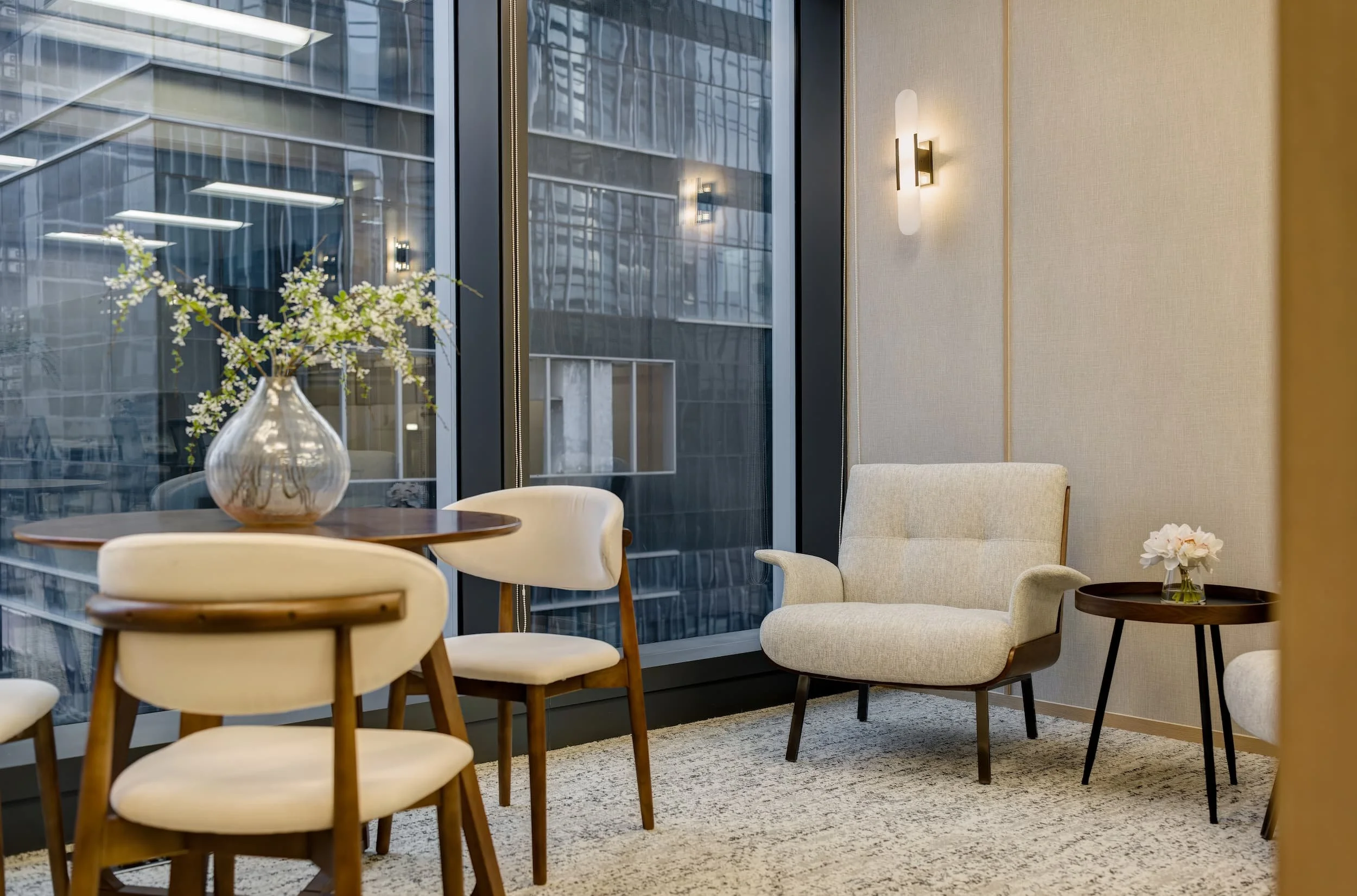 Modern interior space with cream-colored armchair, wooden chairs around a table with a vase of flowers, large window showing city buildings, and wall-mounted light fixtures.