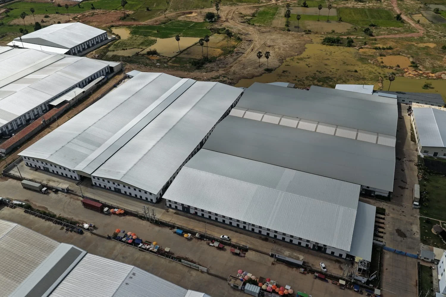 An aerial view of large industrial warehouse buildings with white roofs, surrounded by a parking area and some nearby water bodies, greenery, and trees in the background.