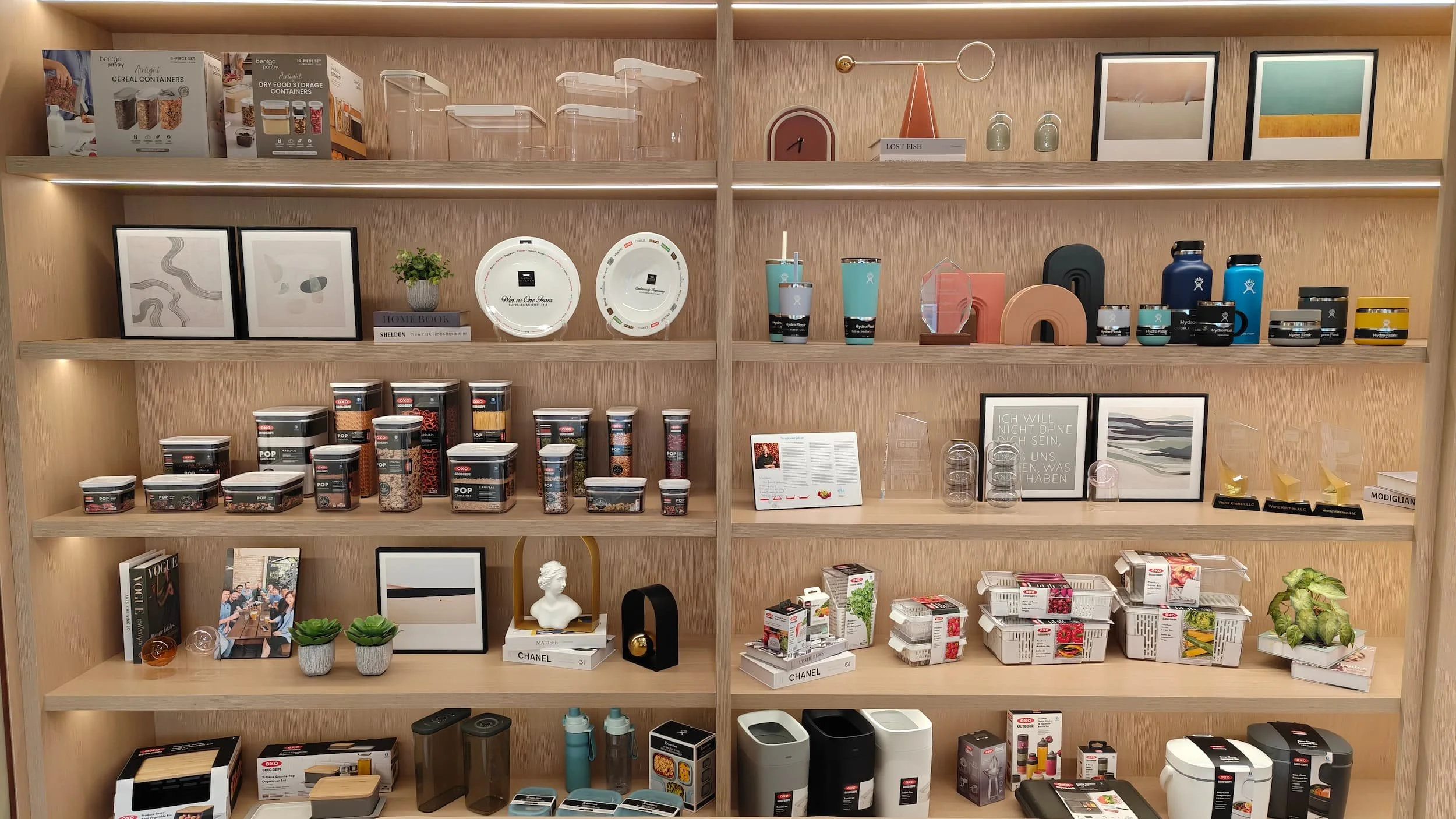 Wooden shelving unit with various kitchen and home decor items, including framed pictures, plants, books, bowls, and storage containers.