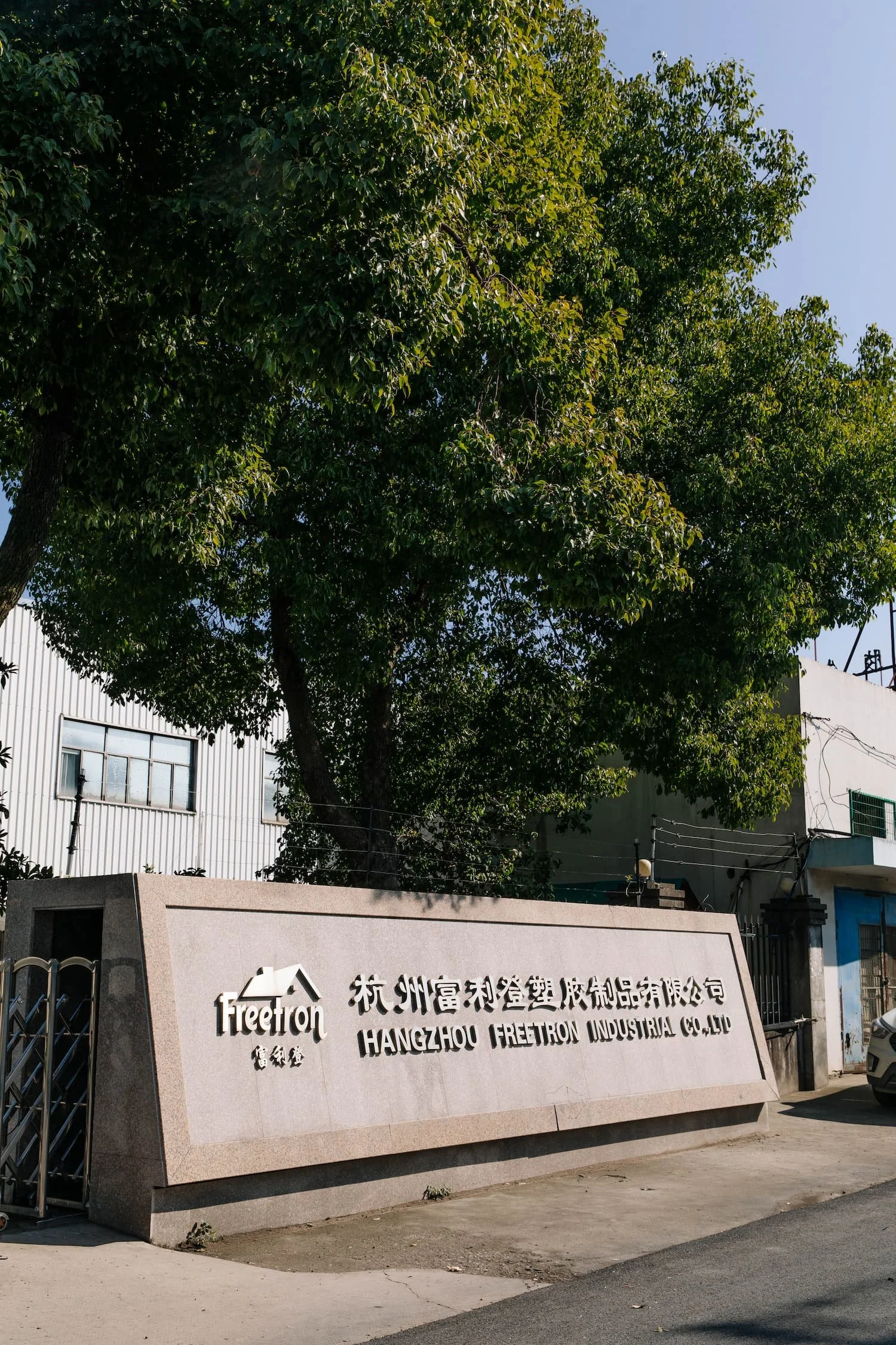 A large stone sign with text in Chinese and English saying "Hangzhou Freetron Industrial Co., Ltd." and a logo with a stylized house, situated in front of a building with white walls and windows, surrounded by a lush green tree, under a clear blue sky.