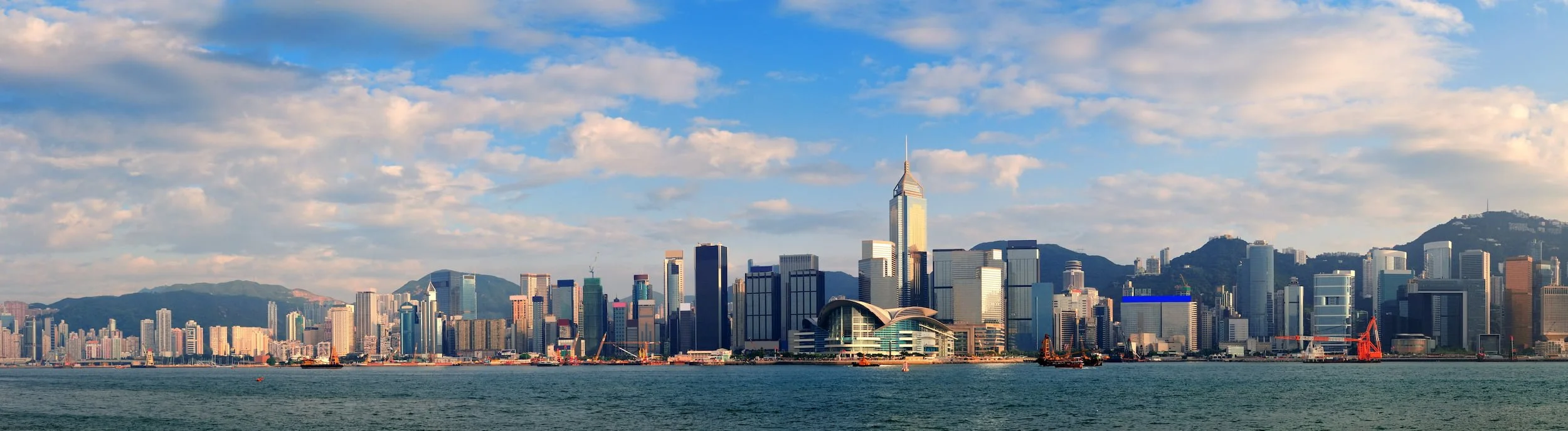 Famous city skyline with skyscrapers along water, featuring a tall, pointed building, mountains in background, with partially cloudy sky.