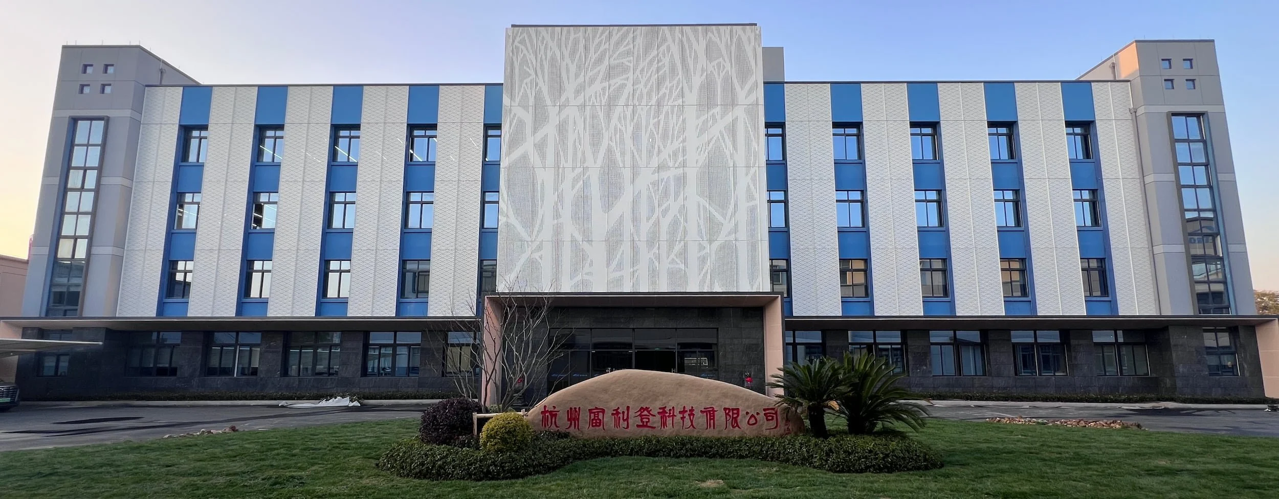 Front view of a modern multi-story building with blue and white panels, large windows, and a decorative panel on the upper facade; there is a lawn with bushes and a sign with red Chinese characters in front.