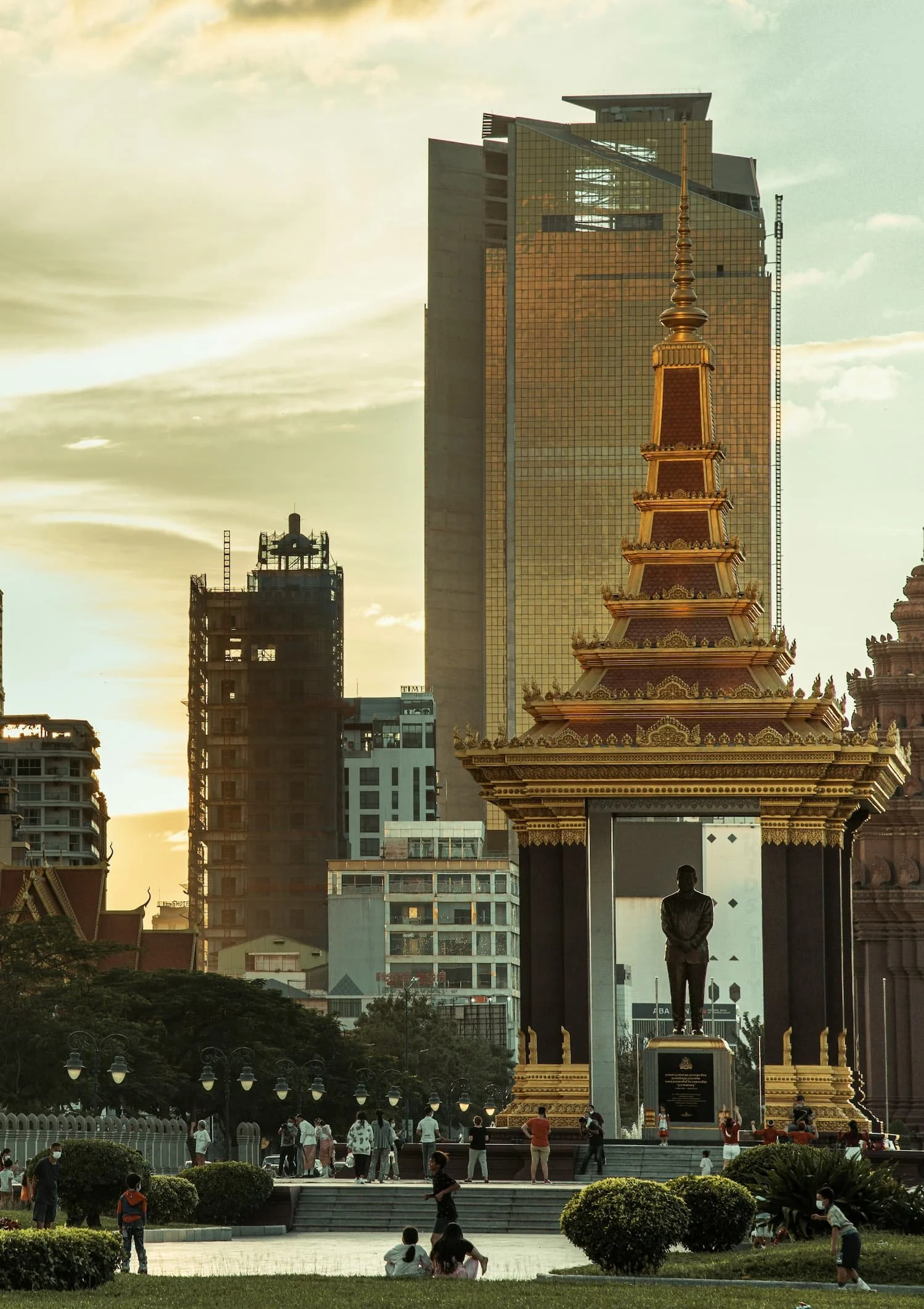 People gather in a park in front of a tall ornate temple with a gold spire, surrounding skyscrapers in the background with a sunset sky.