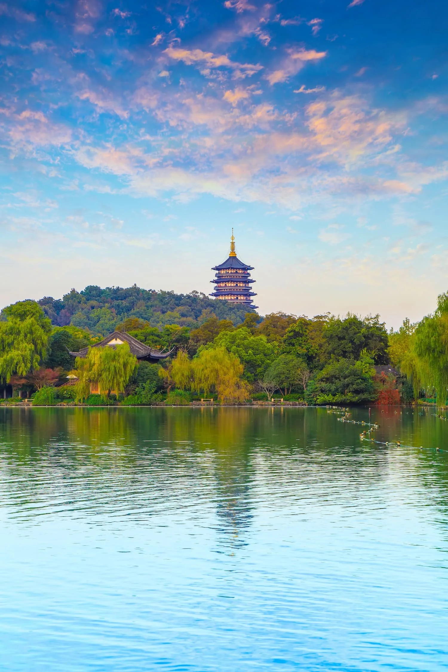 A serene lake with calm water reflecting a sky during sunset, with pastel pink and blue clouds. In the background, a pagoda-style tower is perched on a lush, green hill surrounded by trees and a traditional building with a curved roof.