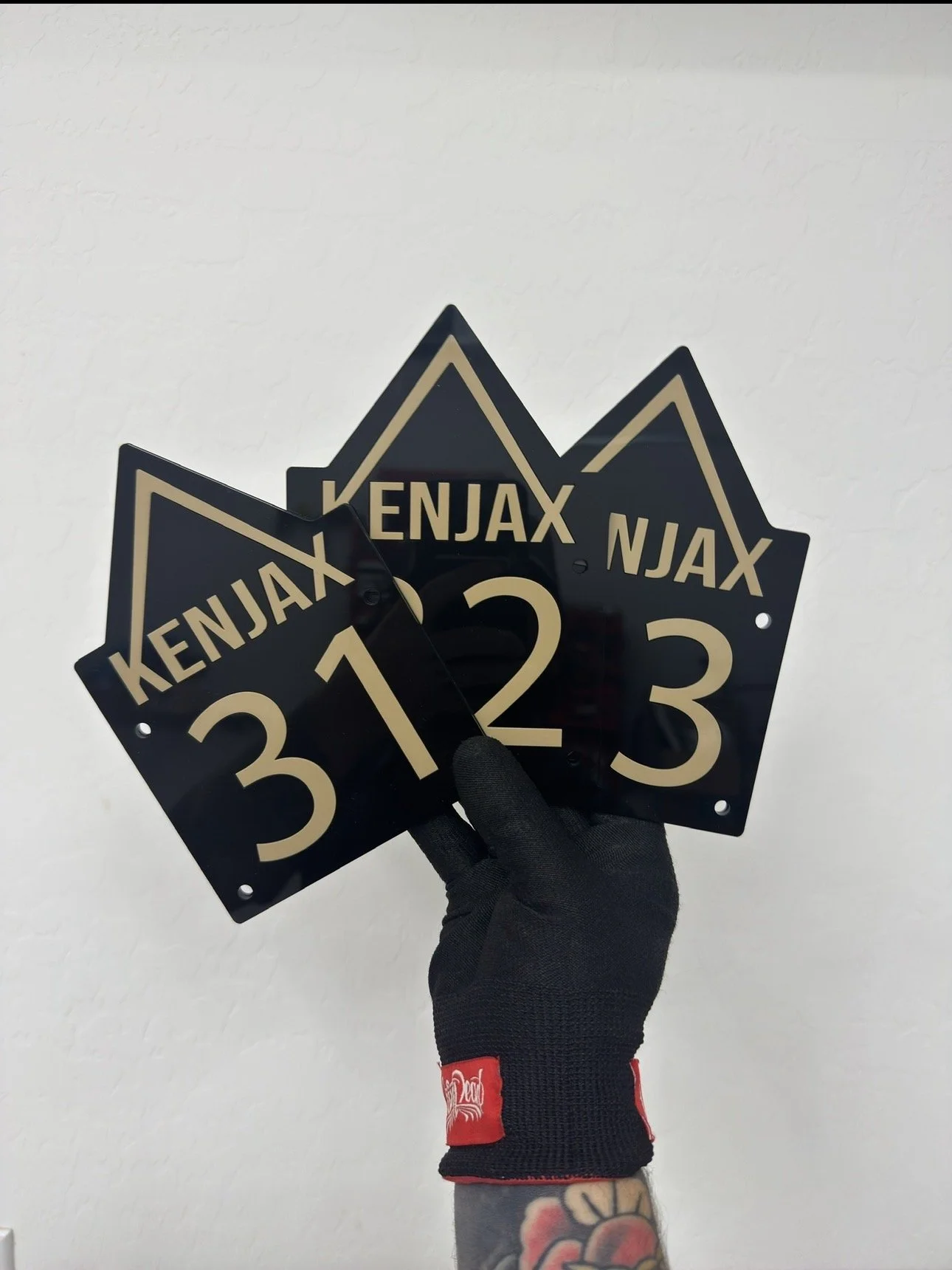 Hand wearing a black glove holding three black numbers with beige lettering: 3, 12, 23, and the word 'NJAX' printed on each, against a plain white wall.