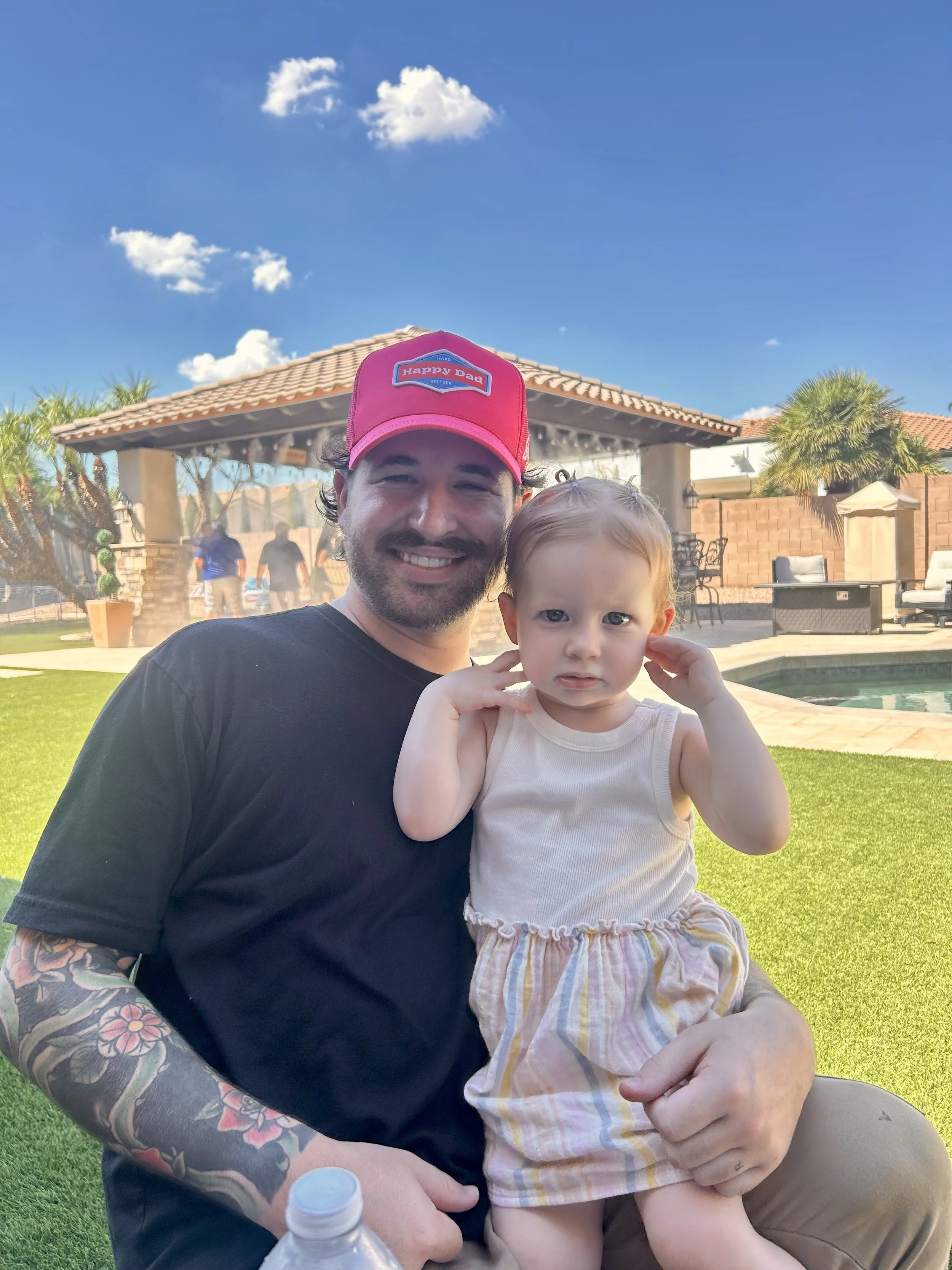 Man wearing a red 'Happy Dad' baseball cap holding a young girl in a cream tank top and striped skirt outdoors in a backyard with a swimming pool, patio furniture, palm trees, and a gazebo under a blue sky with some clouds.