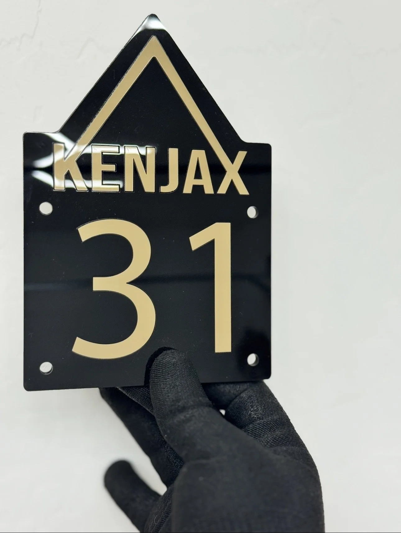 Close-up of a black house-shaped sign with the word "KENJAX" at the top and the number "31" below, being held by a hand wearing a black glove.