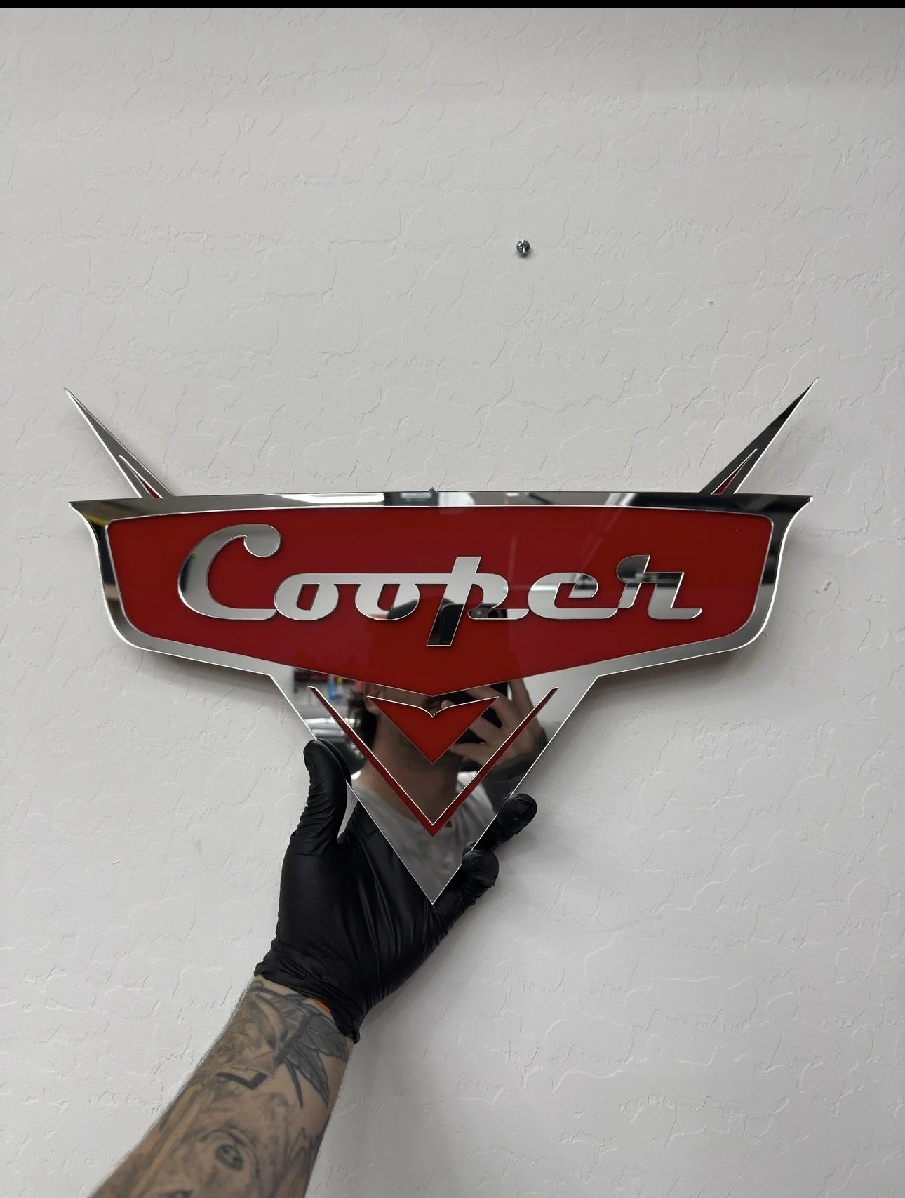 Person with tattooed arm and black glove holding a reflective, chrome and red 'Cougher' logo sign mounted on a wall.