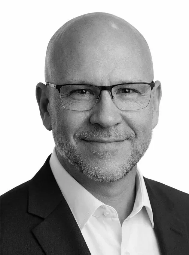 Black-and-white professional headshot of a middle-aged man with glasses, a bald head, and a short beard, wearing a suit and white shirt, smiling.