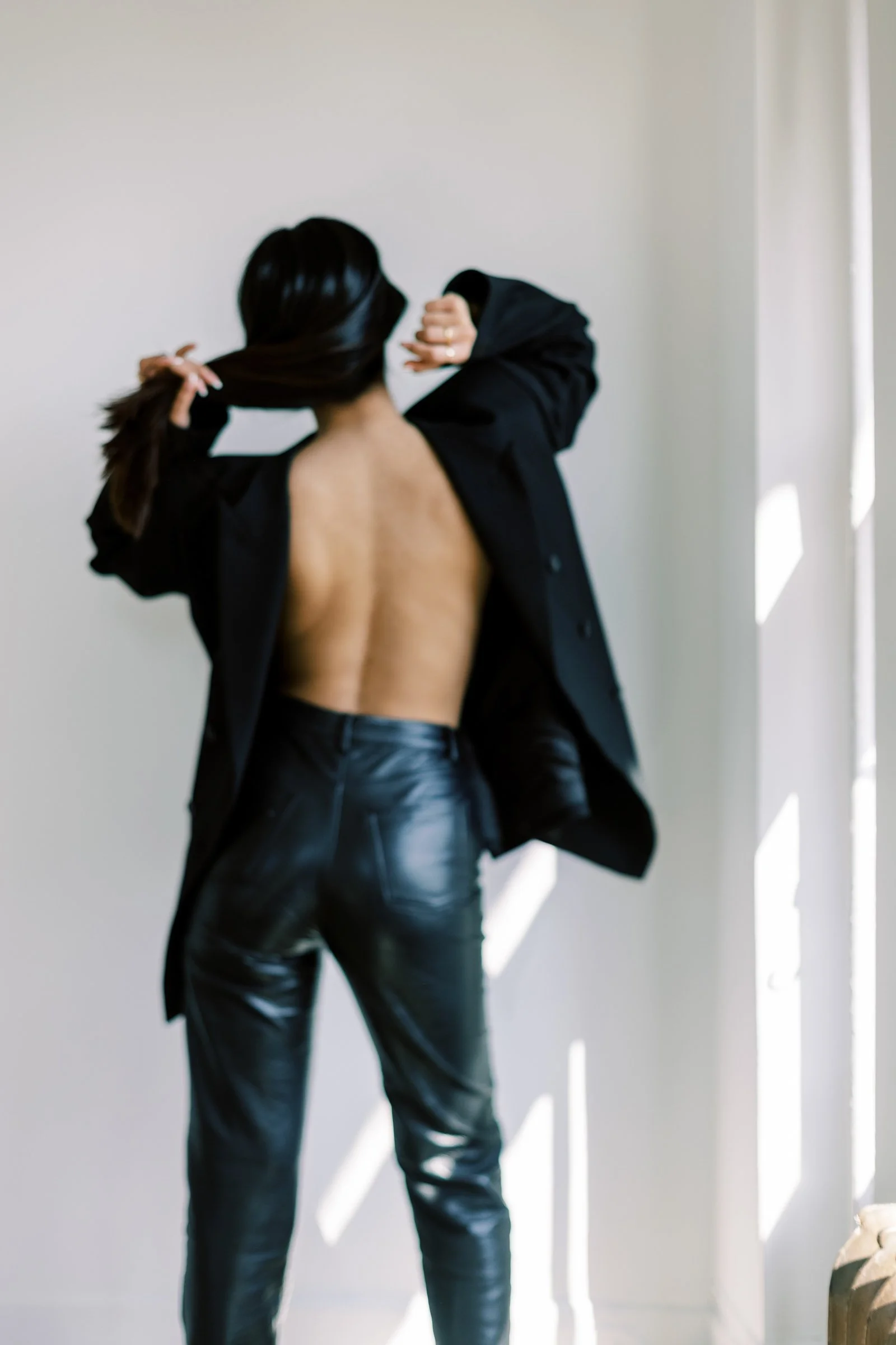 A woman with long dark hair, wearing a black blazer and black leather pants, is standing with her back to the camera, fixing her hair against a light gray wall with sunlight casting shadows.