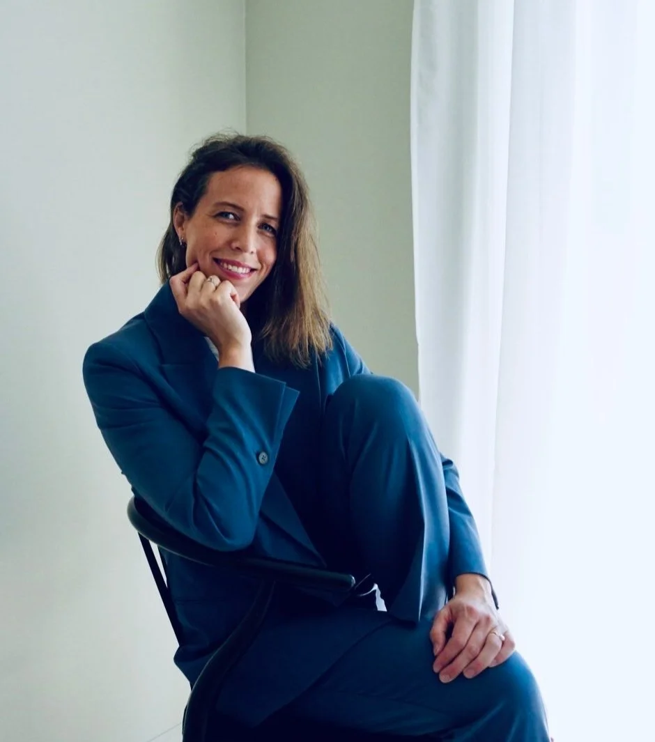 A woman in a blue suit sitting on a chair near a window with white curtains, smiling and resting her chin on her hand.