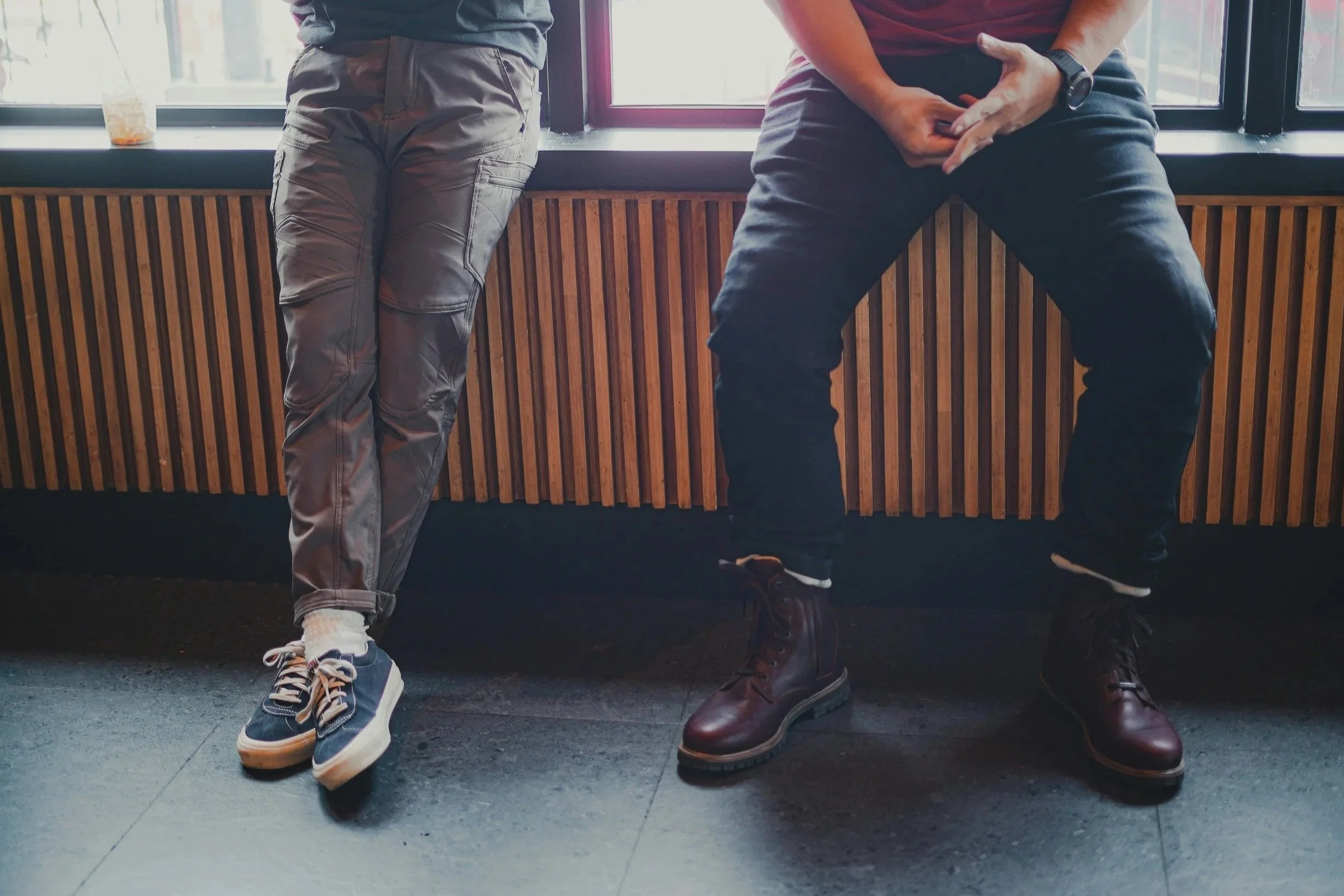 Two men sitting together in conversation, legs and feet visible, in a warm café setting