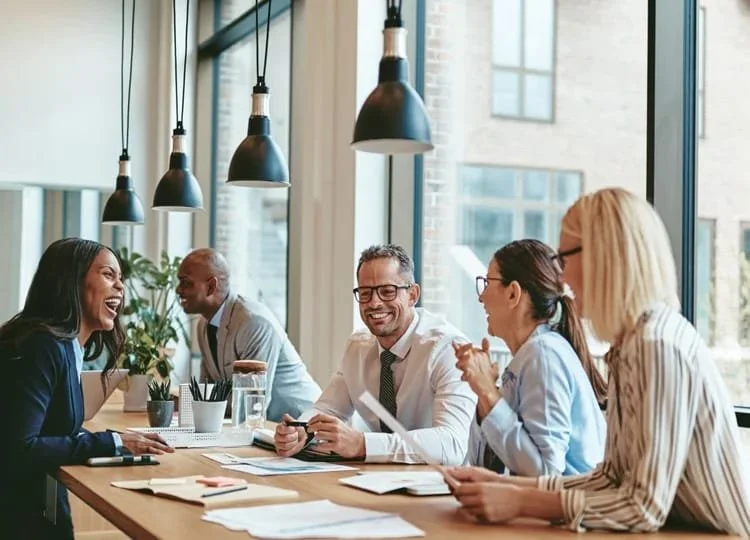 Ein Team von Kollegen lacht gemeinsam während einer Besprechung an einem Holztisch.