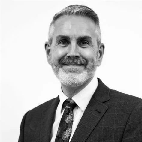 Black and white photo of a smiling middle-aged man with short hair, a beard, dressed in a suit and tie.