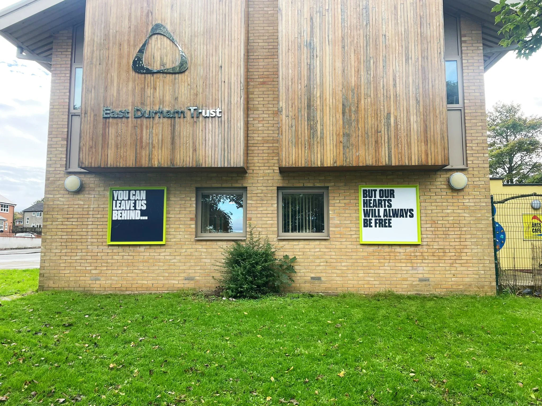 The image shows a two-story brick building with wooden panels on the upper level. There are two large windows on the upper level, and a door with glass windows on the lower level. The building has two signs; the left one says "You can leave us behind..." and the right one says "but our hearts will always be free." There is a small shrub in front of the building and a grassy lawn. A tree with green leaves is partly visible on the upper right corner, and a fence is visible on the lower right side.
