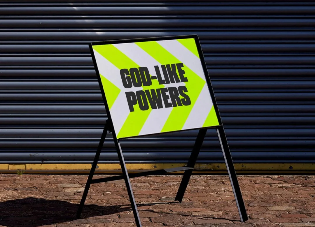 A black A-frame sidewalk sign with a bright yellow and white background and bold black text that reads "GOD-LIKE POWERS," standing on a brick sidewalk in front of a metal corrugated garage door.
