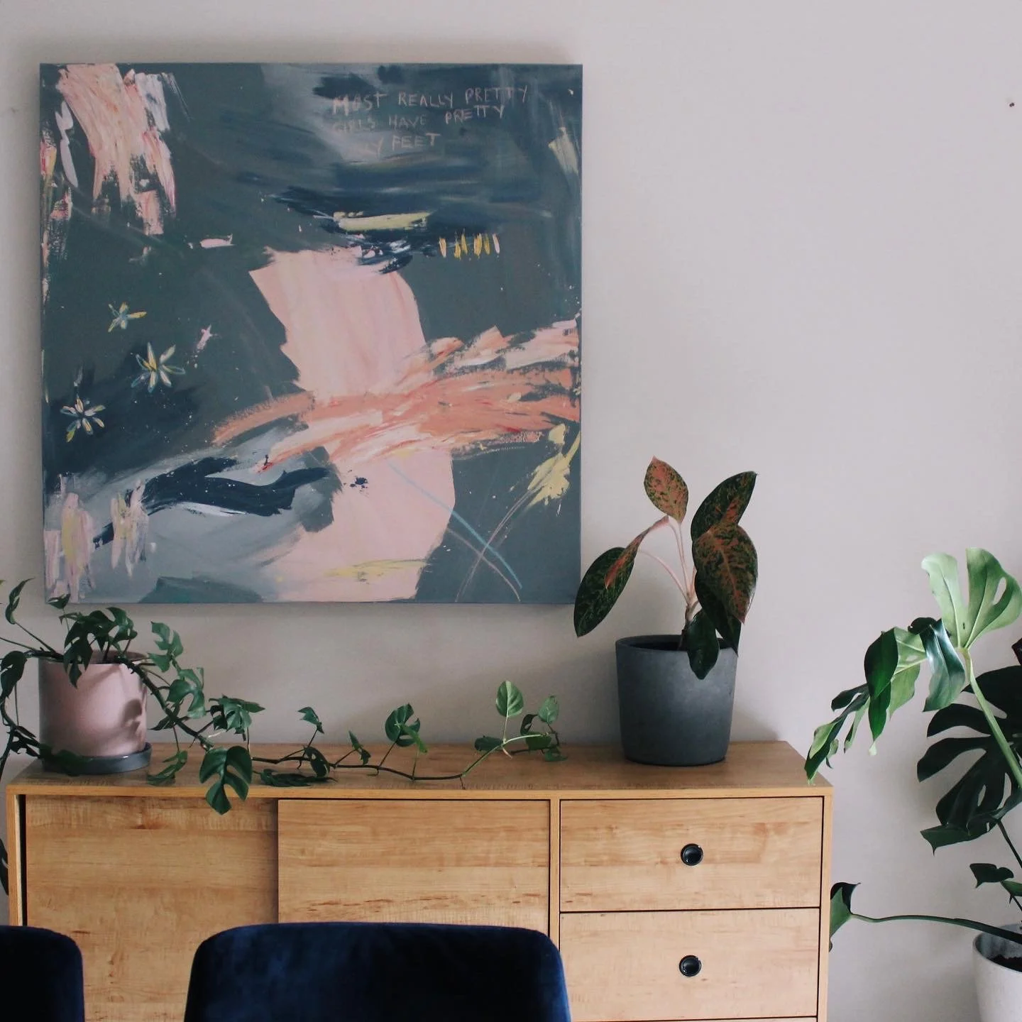 A modern interior with a wooden sideboard topped with potted plants. An abstract painting in shades of blue, pink, and white hangs on the wall behind. The plants include a vine with green leaves cascading over the sideboard and a larger plant with re