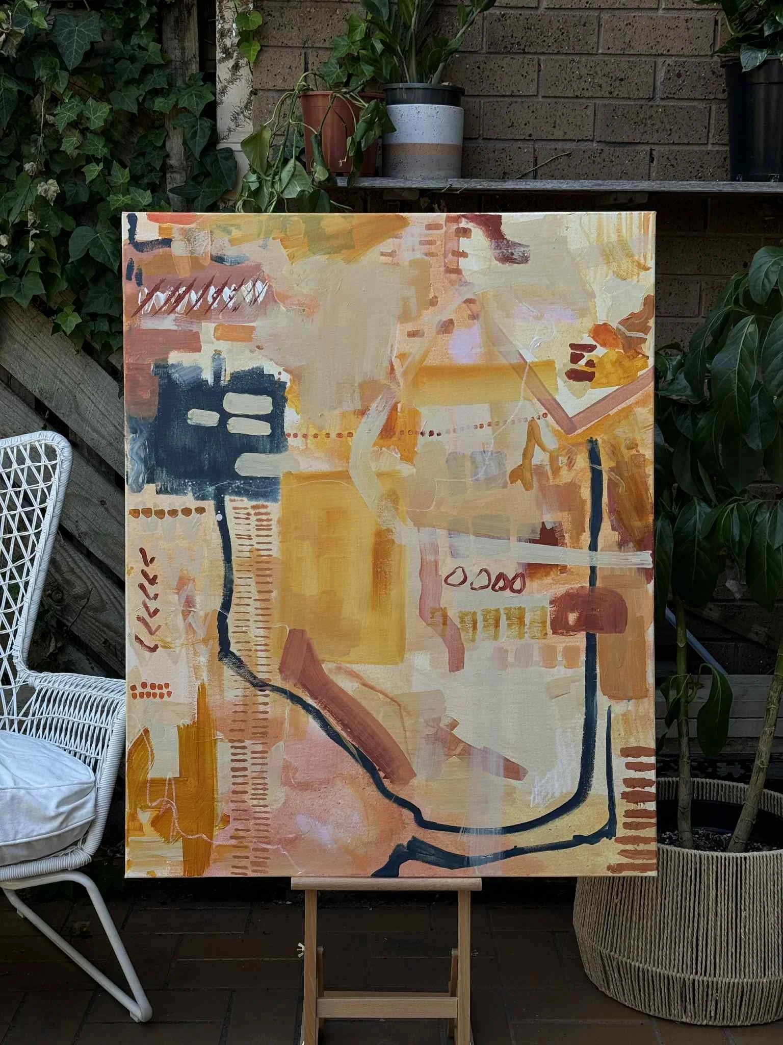 An abstract painting with warm tones, featuring various brushstrokes, patterns, and shapes, mounted on a wooden easel outdoors.