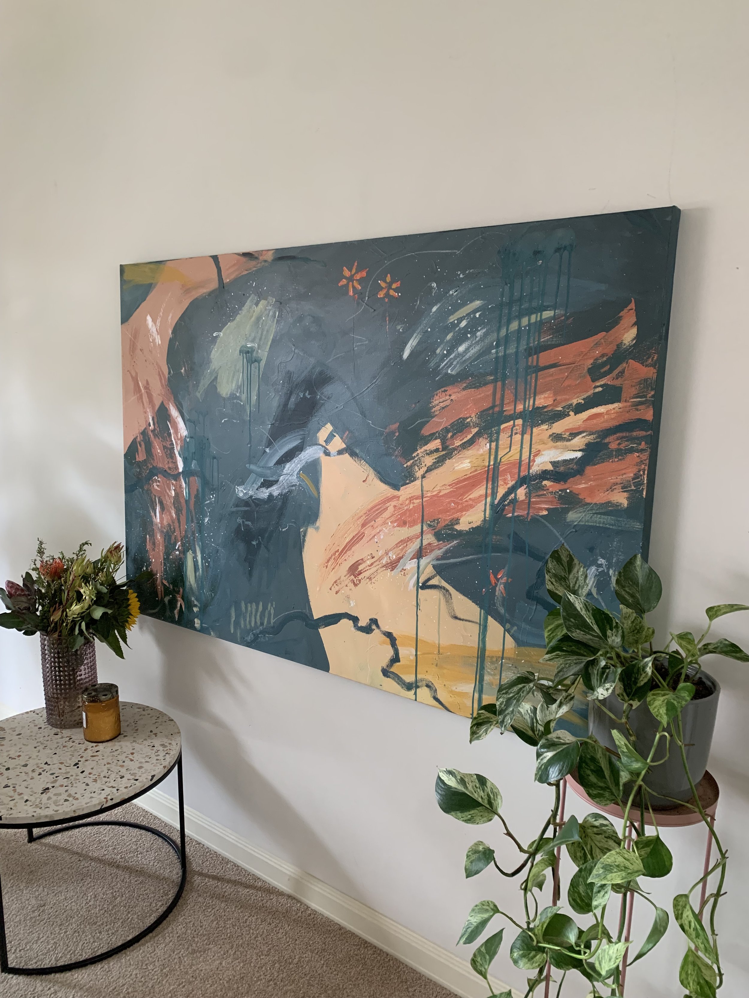 A corner of a room with an abstract painting on a white wall. Below the painting is a small round table with a terrazzo surface, holding a brown vase with a colorful flower arrangement and a small amber candle. To the right, there is a leafy green pl