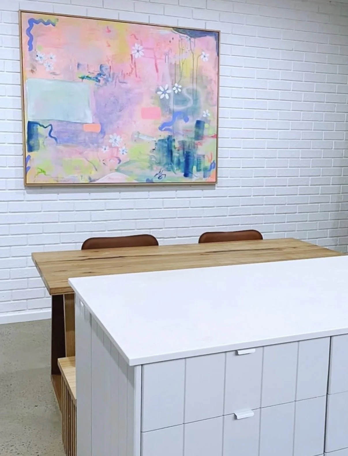 A white cabinet with drawers in front of a wooden table with chairs, and an abstract pastel-colored painting on a white brick wall.