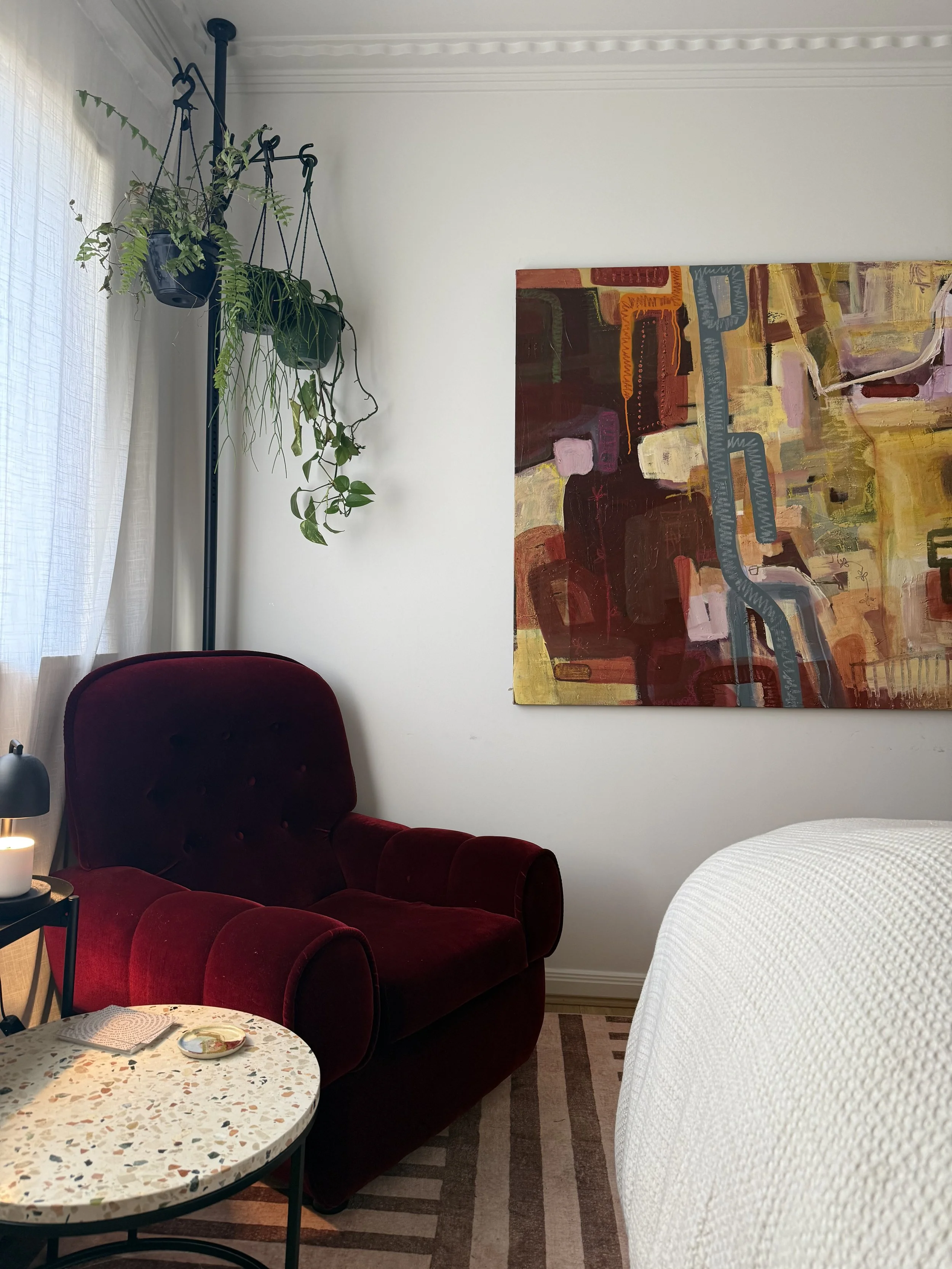 A cozy living room with a red velvet armchair, a small table with a plate, a framed picture on a side table, a hanging plant, a large abstract painting on the wall, and part of a white bed covering a portion of the right side.