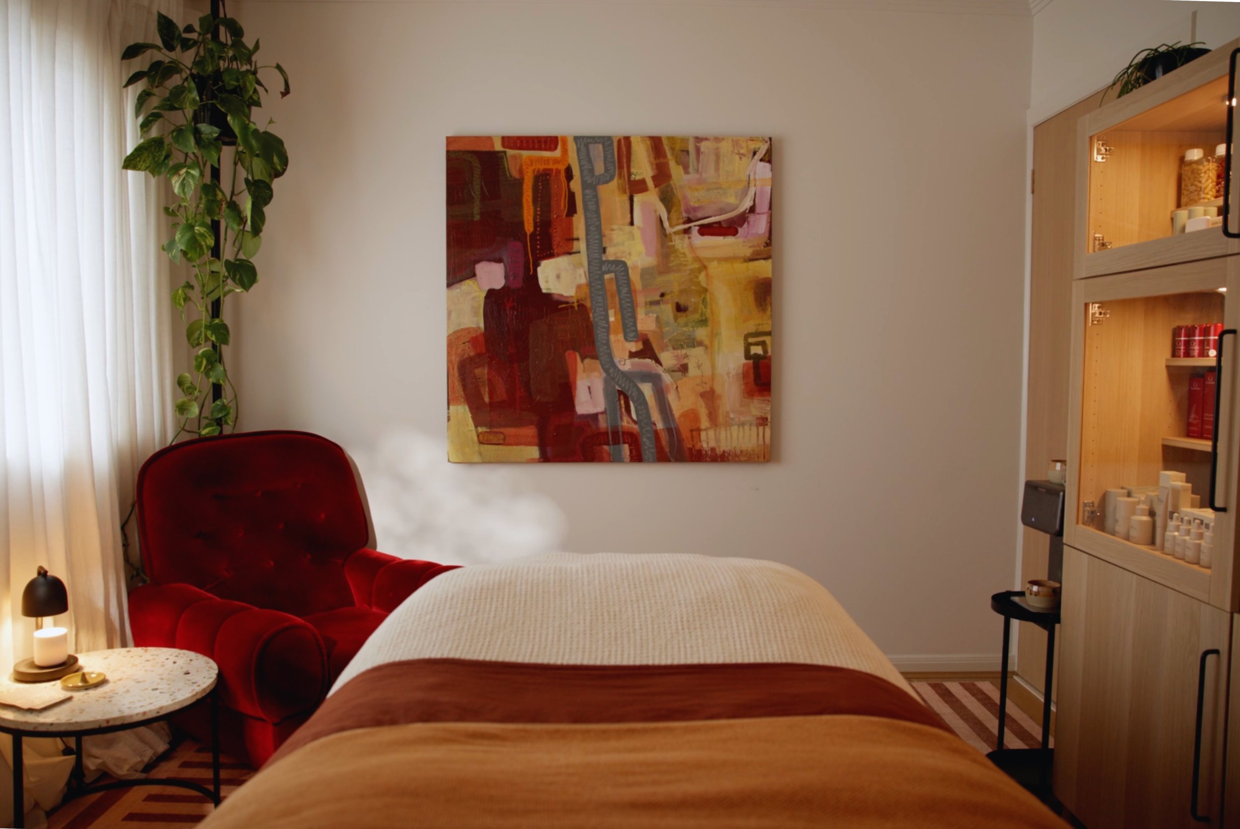 A cozy living room corner with a red velvet armchair, a white lamp, a small round table, and a large abstract painting on the wall. Light filters through white curtains on the left, and a wooden shelf with decorative items and books is on the right.