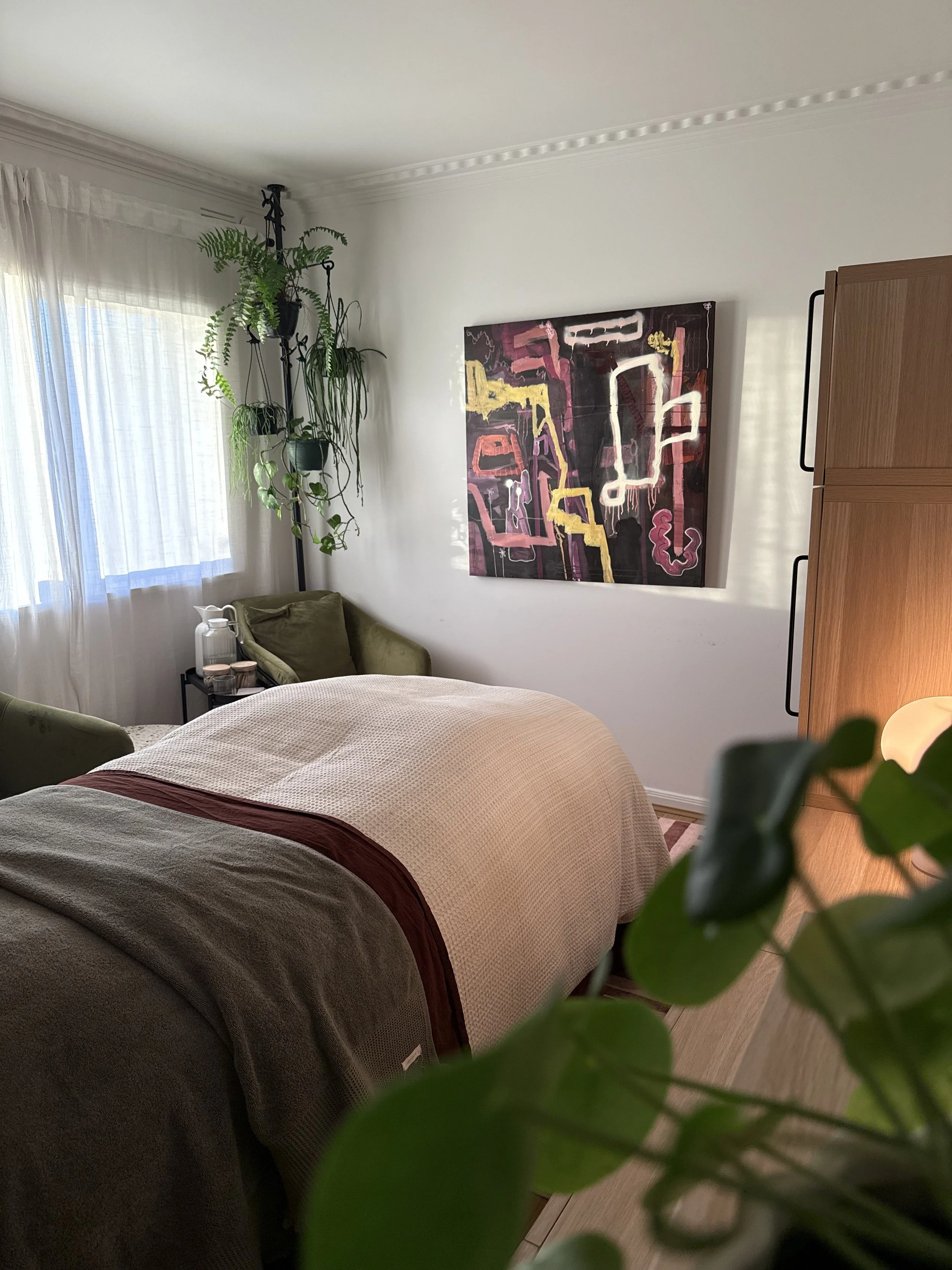A cozy bedroom with a bed covered in a beige blanket, a green armchair, a side table with candles, a tall houseplant with hanging ferns, a window with sheer curtains, a colorful abstract painting on the wall, and a wooden wardrobe.