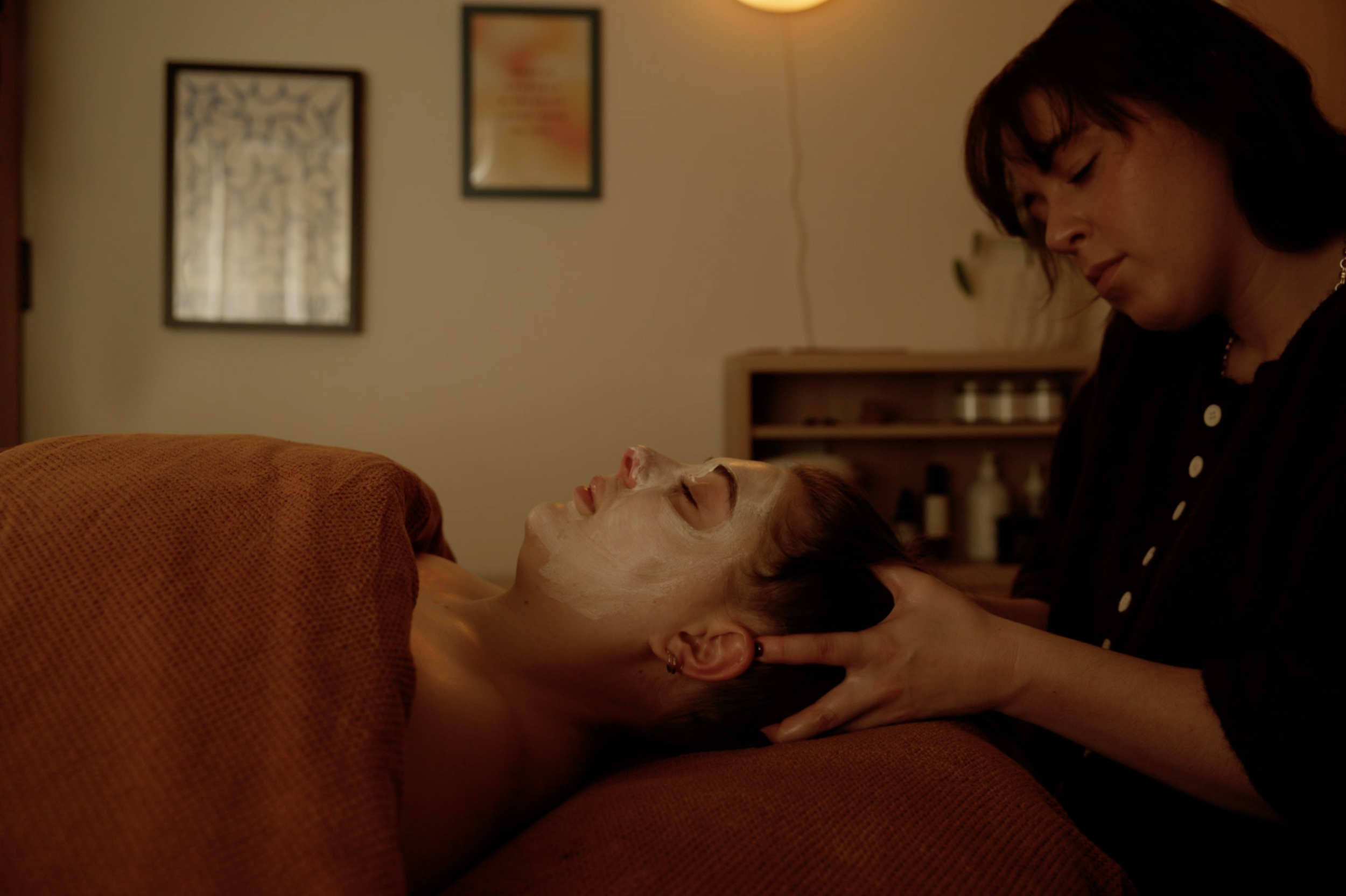 A woman lying on an orange couch with her face partially covered in a facial mask, receiving a head massage from another woman.