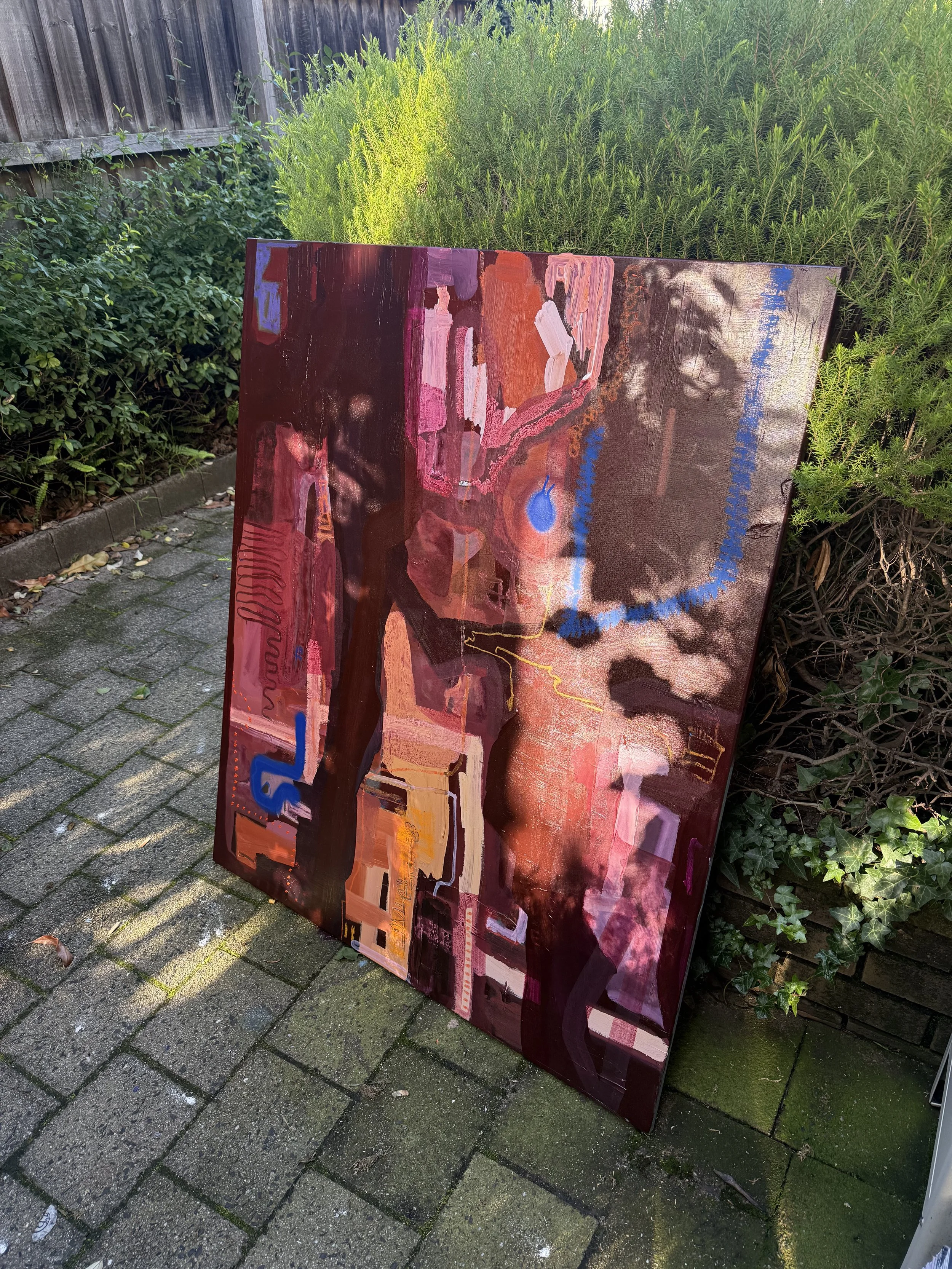 A colorful abstract painting leaning against a bush and a wooden fence outdoors on a paved surface.