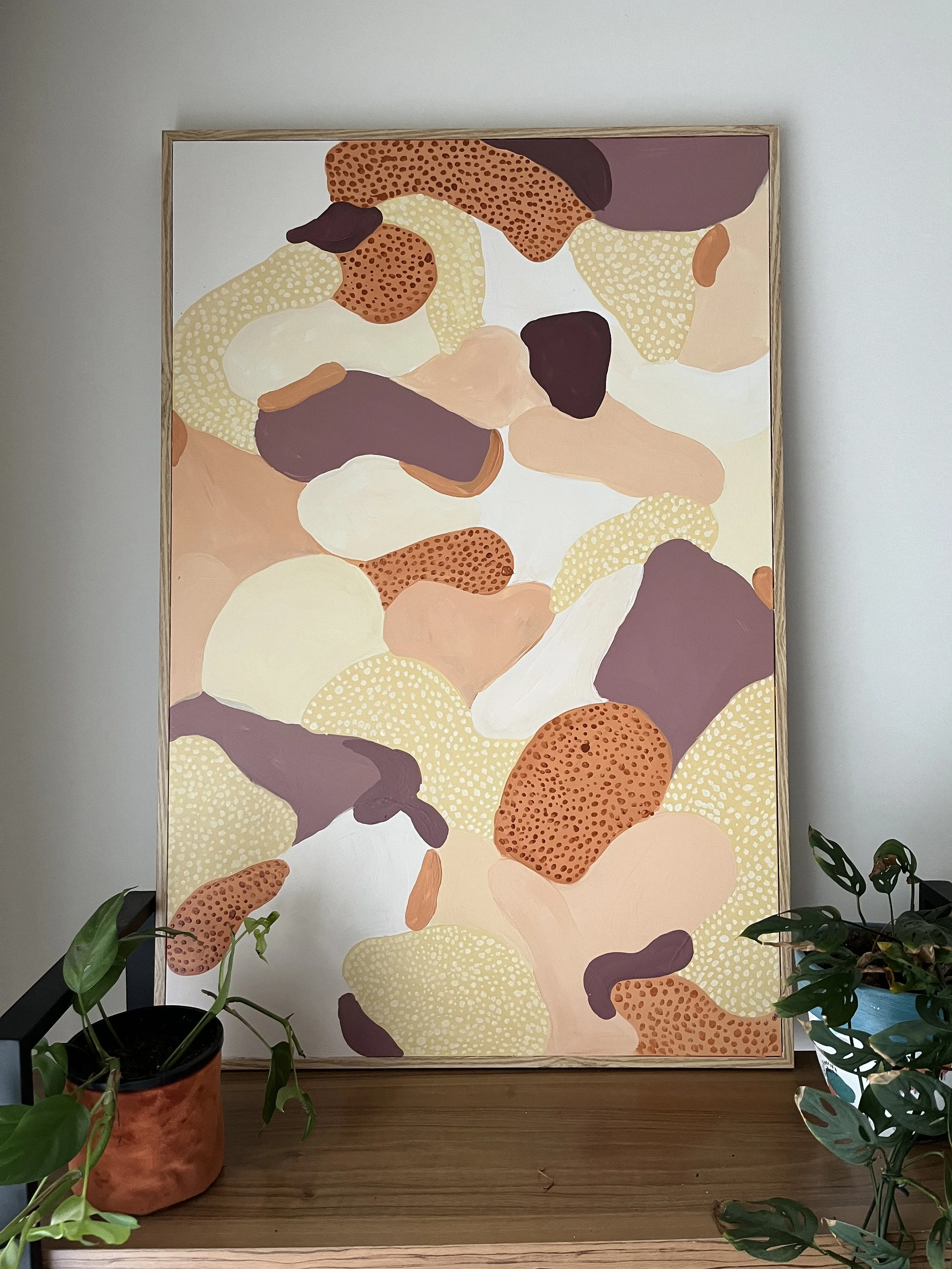 An abstract painting with organic shapes and earthy colors, framed and leaning against a wall in a room with potted plants on a wooden surface.