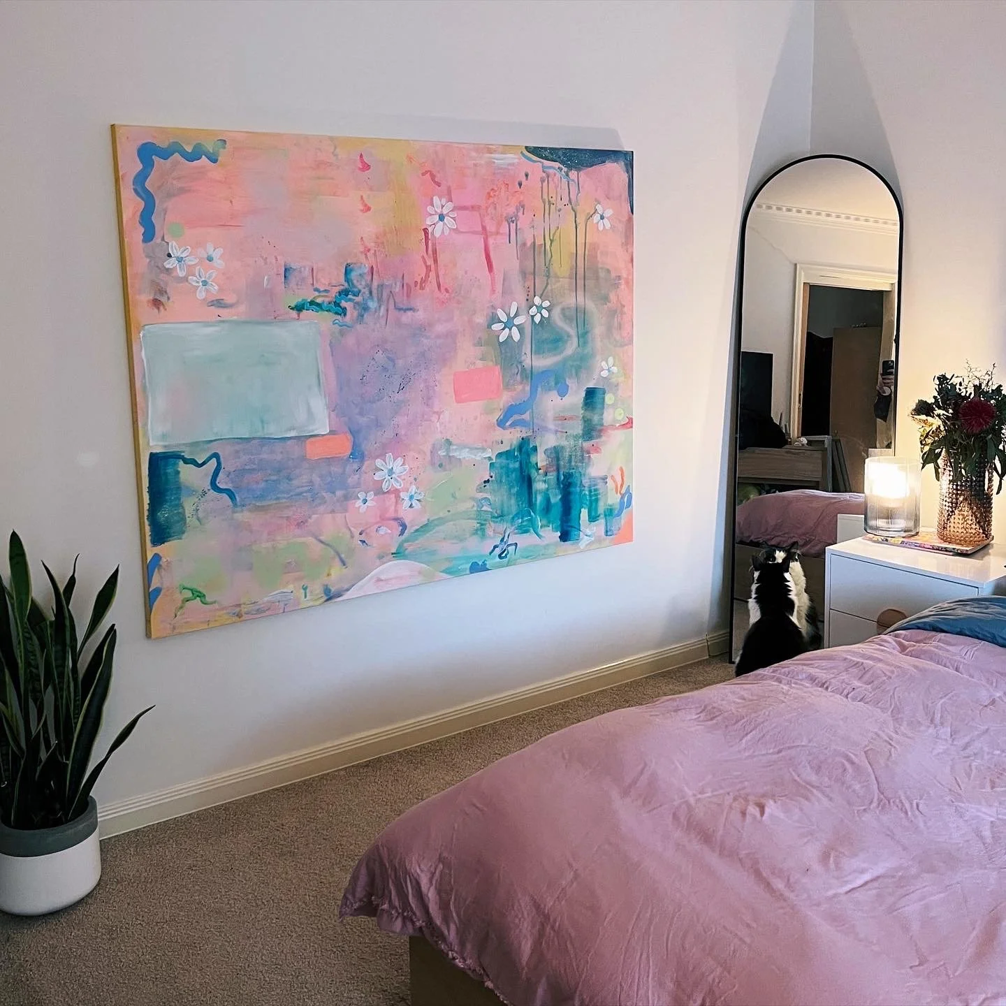 A bedroom with a pastel pink bedspread, a large abstract painting on the wall with pink, blue, yellow, and green colors and floral patterns, a full-length mirror, a white bedside table with a candle and a vase with flowers, a black and white cat sitt