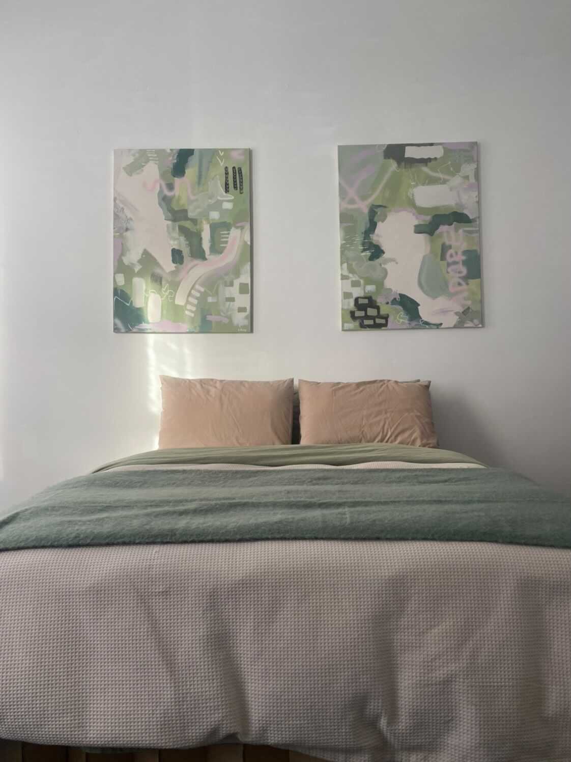 A neatly made bed with two beige pillows against a white wall. Above the bed are two abstract landscape paintings with shades of green, gray, and pink. The bed is covered with a textured beige bedspread and a layered green throw blanket.
