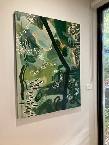 Abstract painting with various green, white, black, and pink shapes and patterns hanging on a white wall near a window.