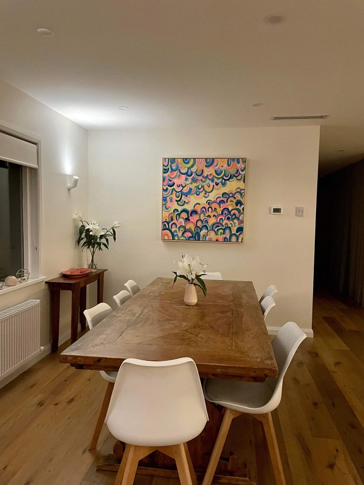 A modern dining room with a large wooden table surrounded by white chairs. A vase with white lilies is on the table. There is a smaller wooden console with plants and pink dishes near the window. The wall features a colorful abstract painting, and th